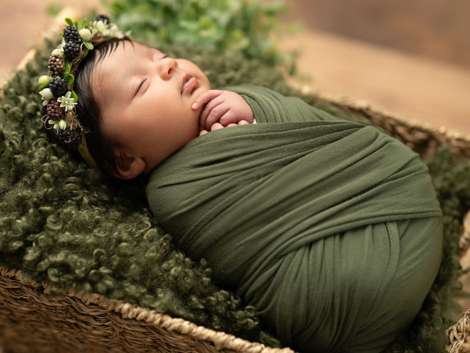 Sleeping newborn wrapped in olive green fabric lying in rustic basket with floral crown and moss bedding.