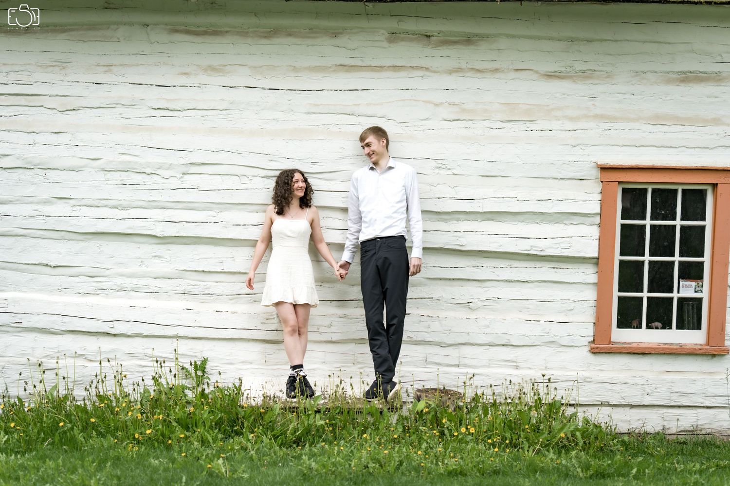 Couple poses casually against white rustic wall with window in countryside setting.