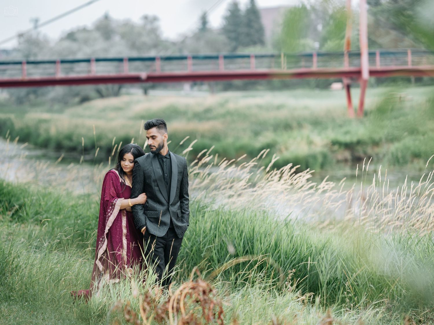 Romantic couple in formal attire standing in tall grass near a red bridge during sunset.