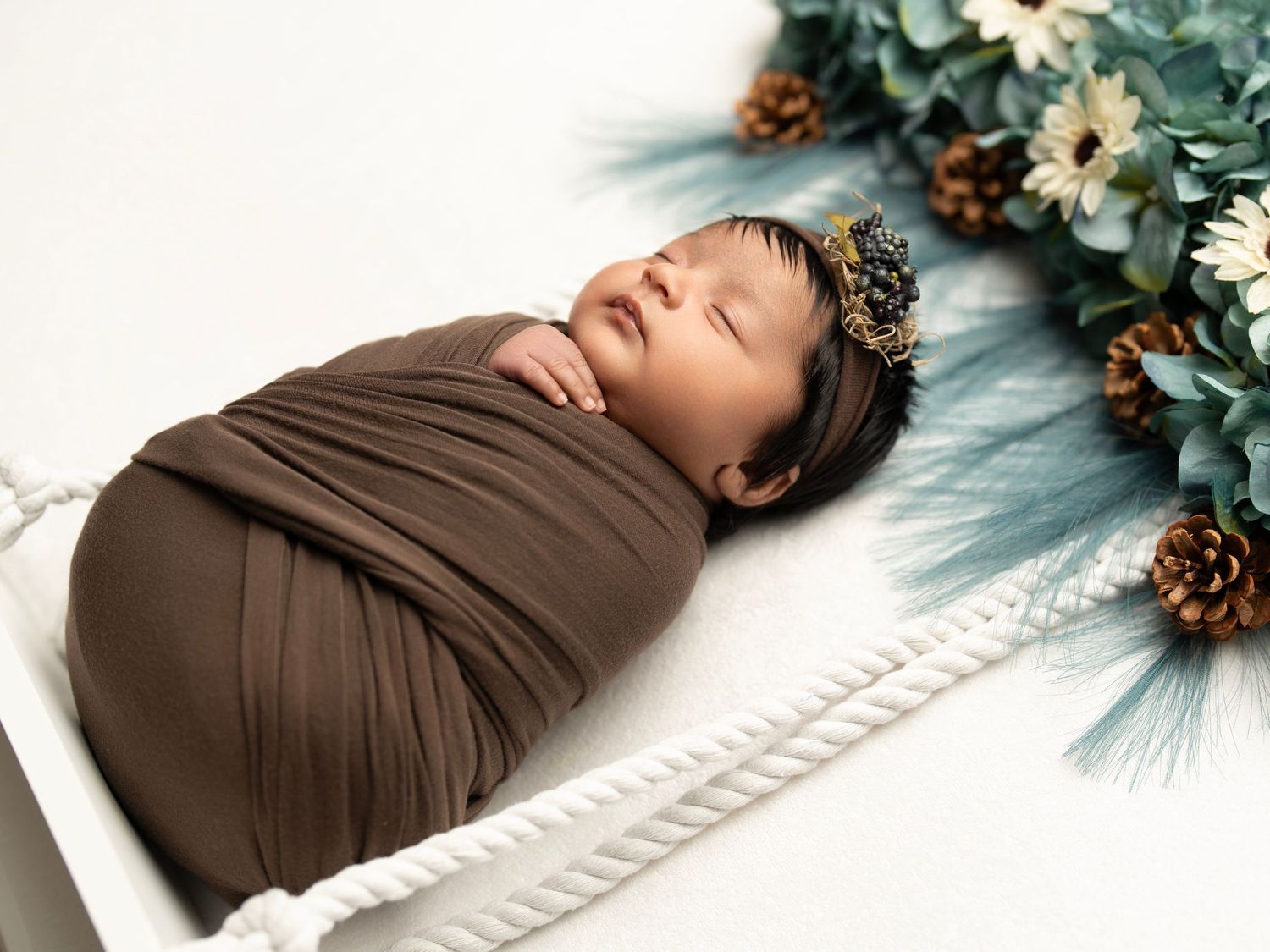 Swaddled newborn baby sleeping on white rope basket with pine branches and floral decor.