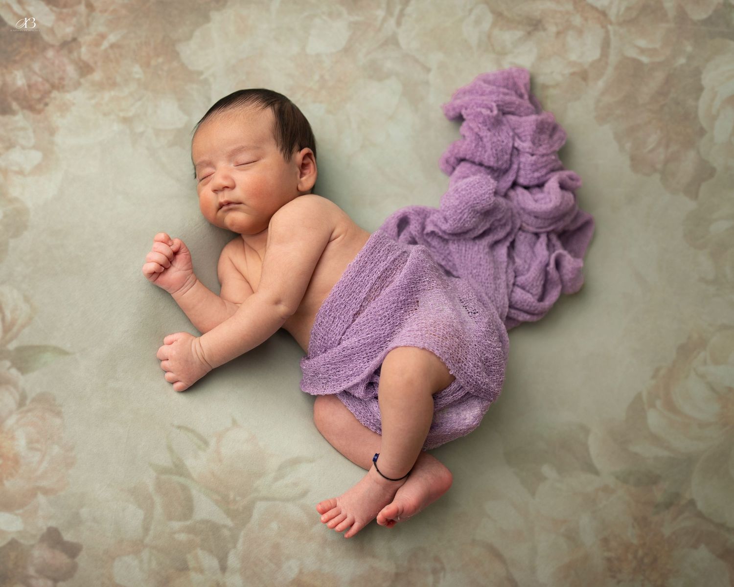 Sleeping newborn posed in lavender knit wrap on textured beige backdrop.