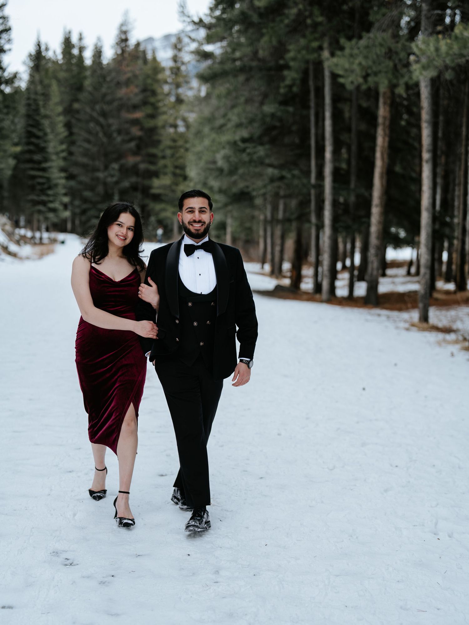 Elegant couple in formal attire walking through a snowy forest path.