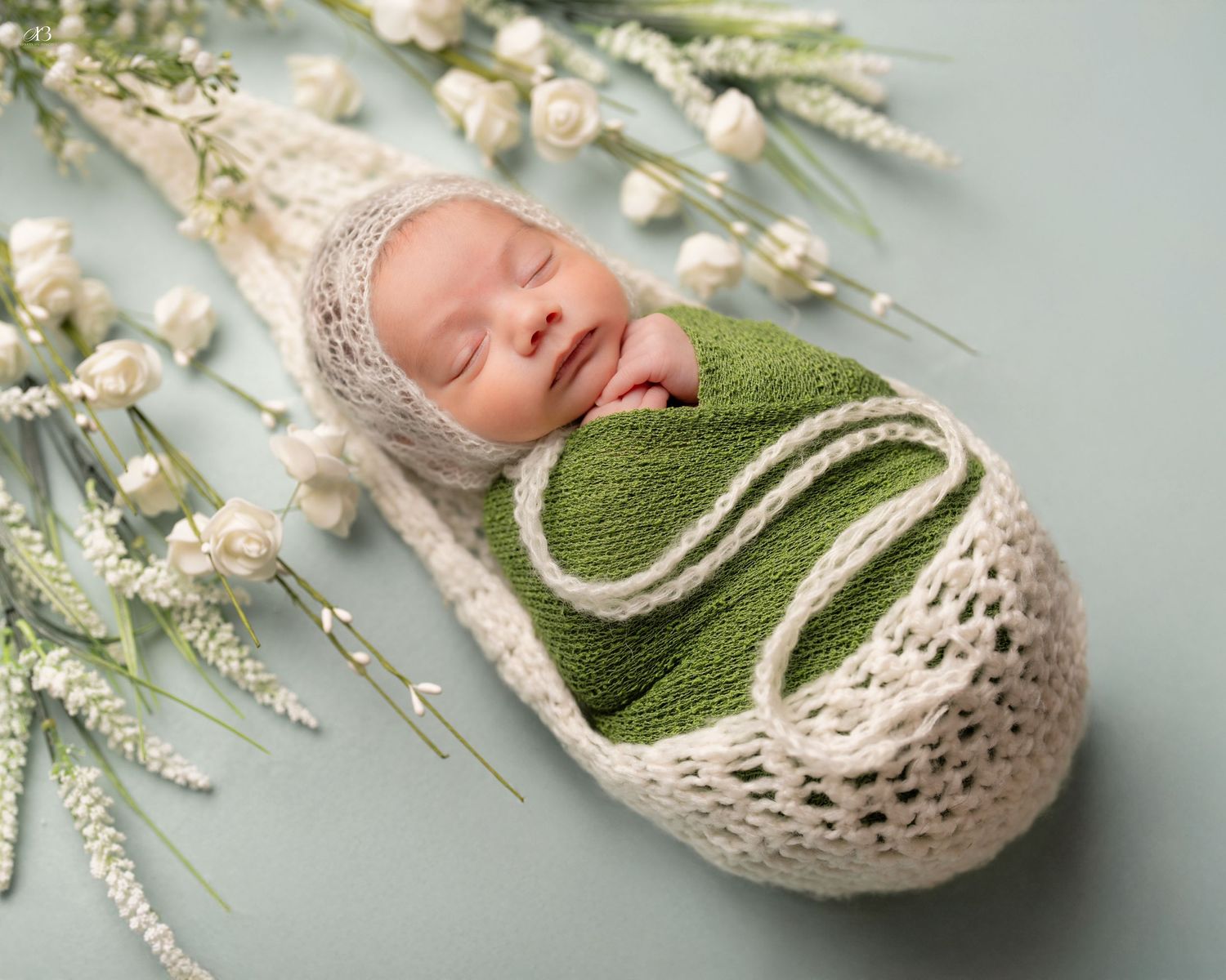 Newborn in green knit wrap and beige bonnet with white florals and branches.