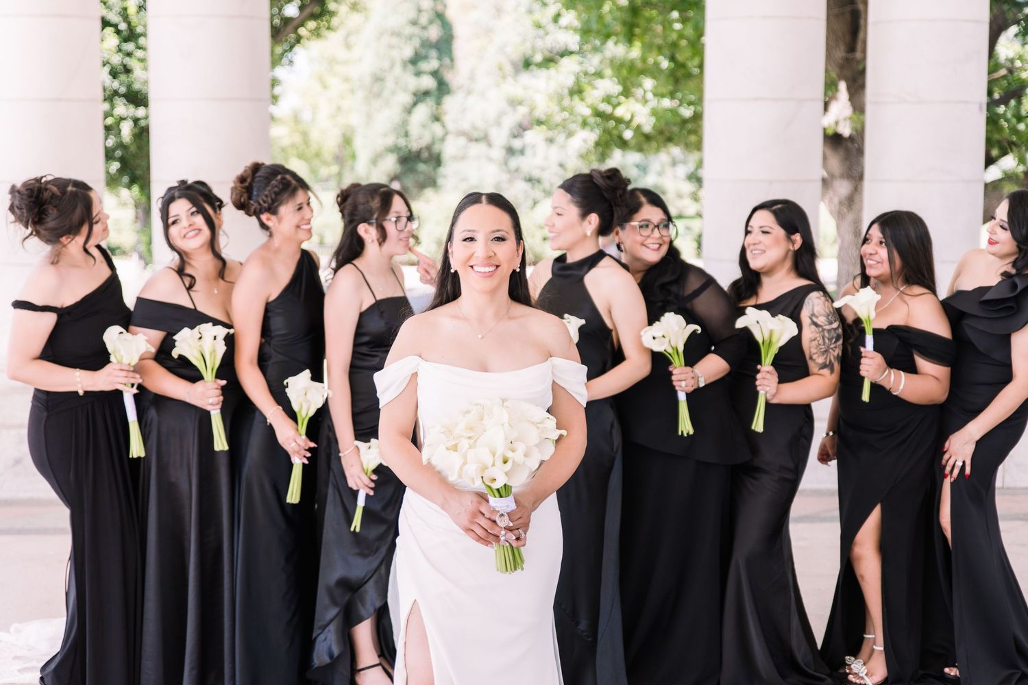 Bride in white gown poses with bridesmaids in black dresses holding white calla lily bouquets outdoors.