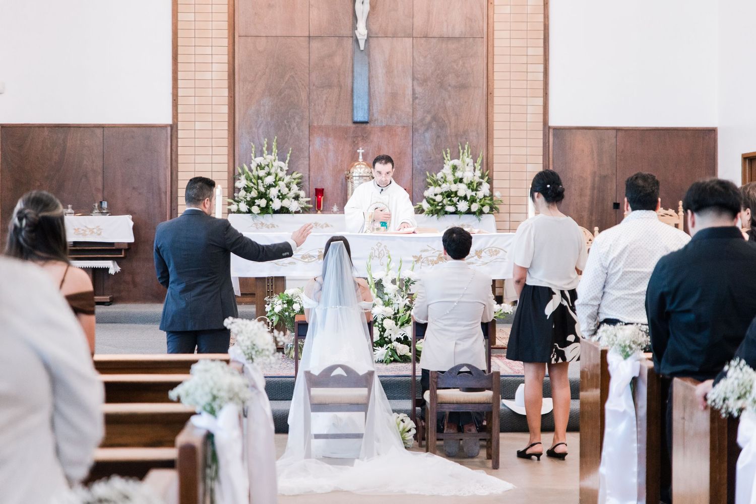 Priest officiates Catholic wedding ceremony as bride and groom kneel at the decorated church altar.