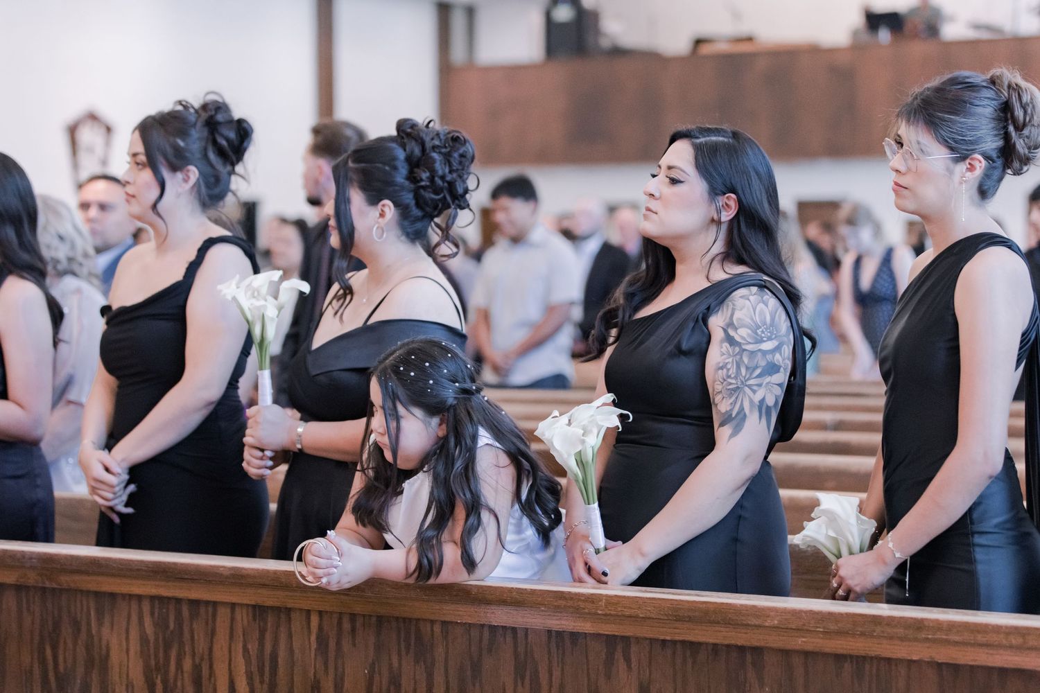 Bridesmaids in black dresses holding white calla lilies stand in church pews during wedding ceremony.