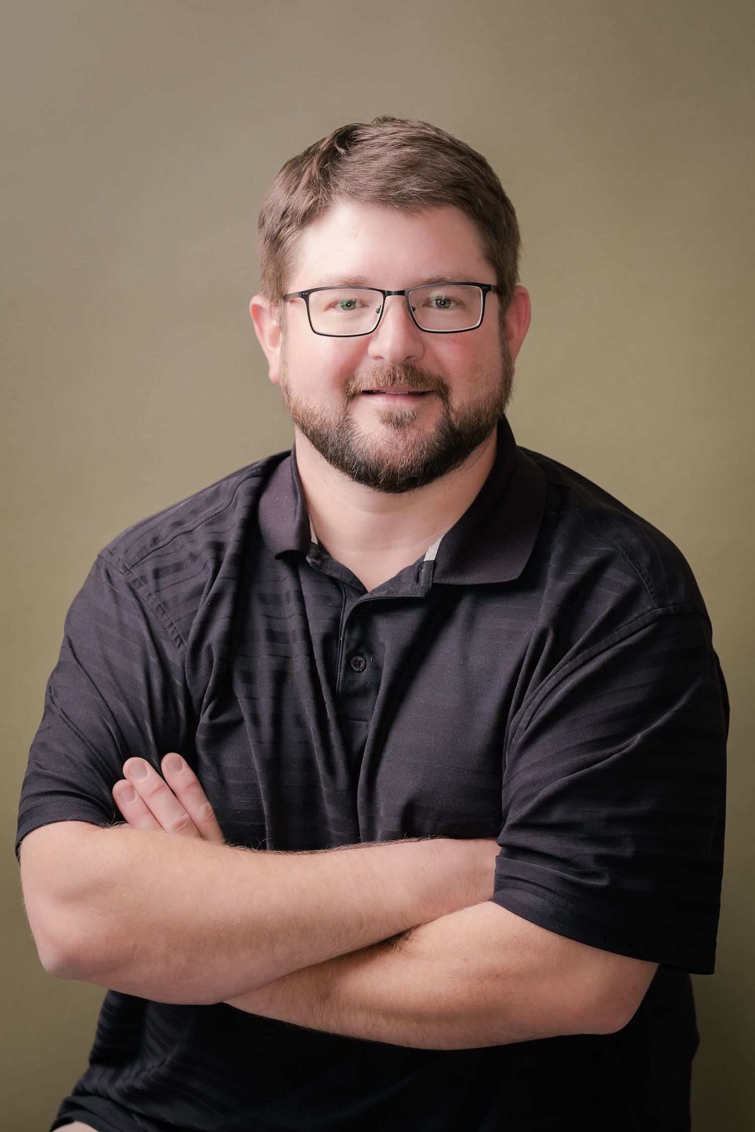 Smiling man with glasses and a trimmed beard wearing a black polo shirt, posing with arms crossed against an olive background.