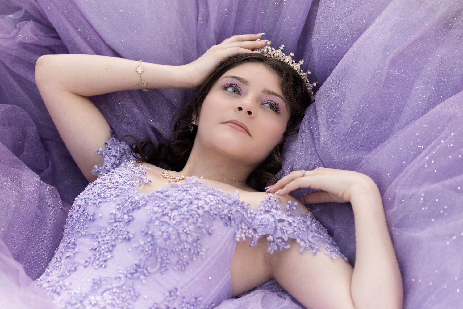 Young woman in lavender gown with floral lace bodice and tiara poses regally against purple tulle fabric.