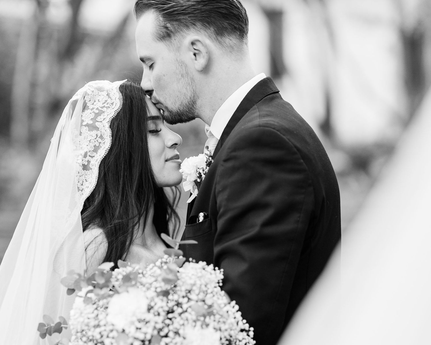 Black and white wedding photo of a groom in a suit kissing his bride's forehead as she wears a lace veil and holds a bouquet.