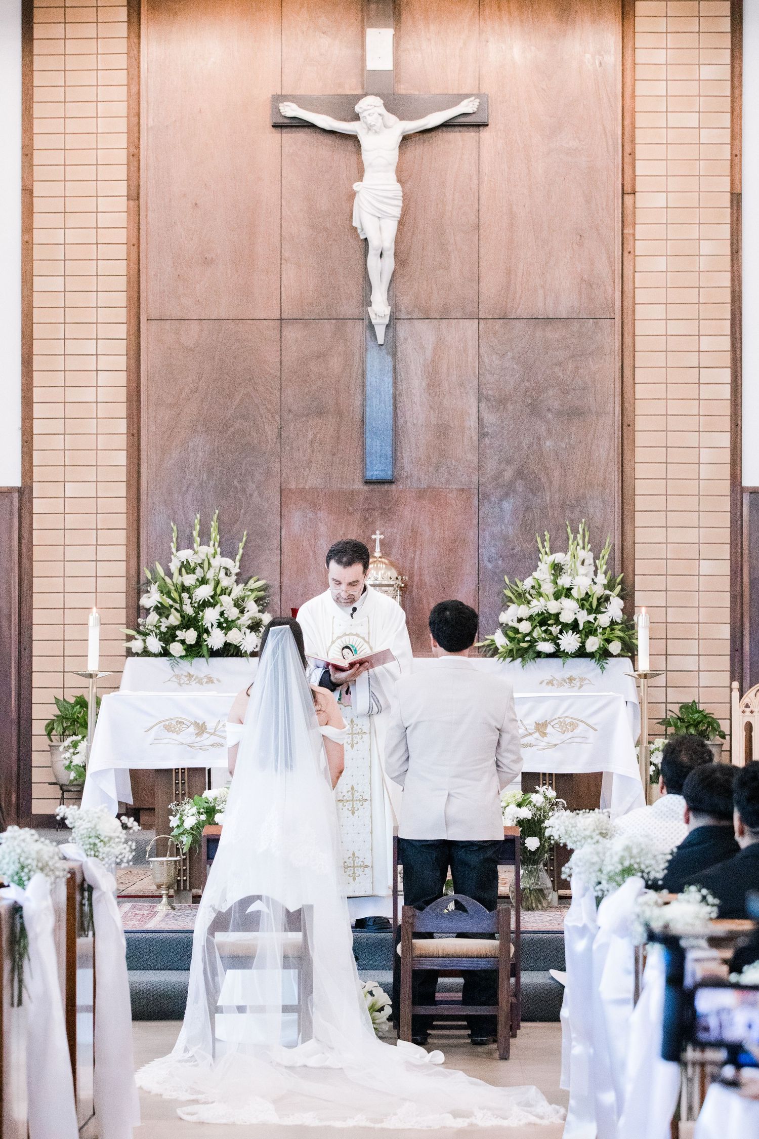 Bride and groom exchange vows at Catholic church altar with crucifix and white floral arrangements.