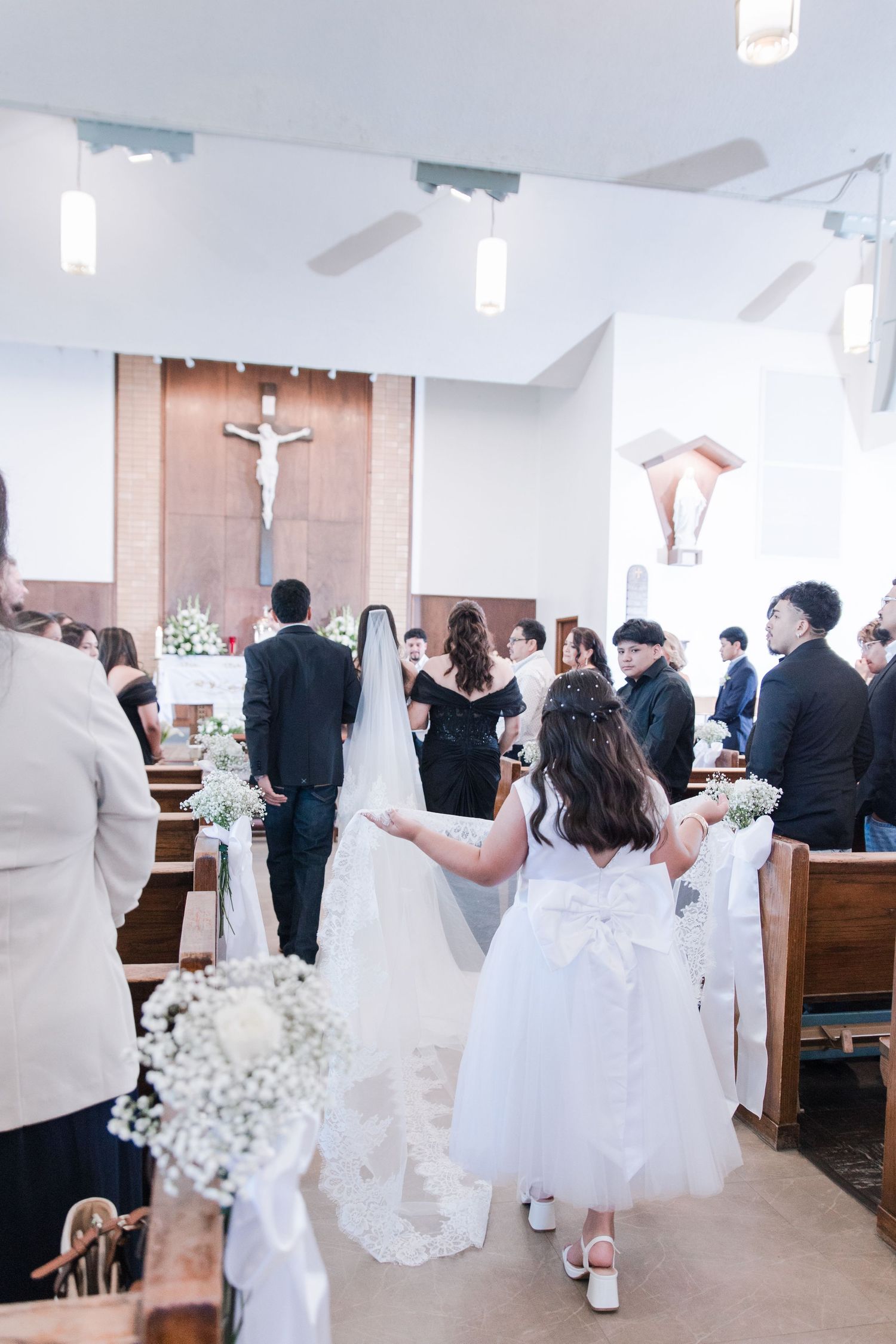 Flower girl carries bride's veil down church aisle during Catholic wedding ceremony with guests seated.
