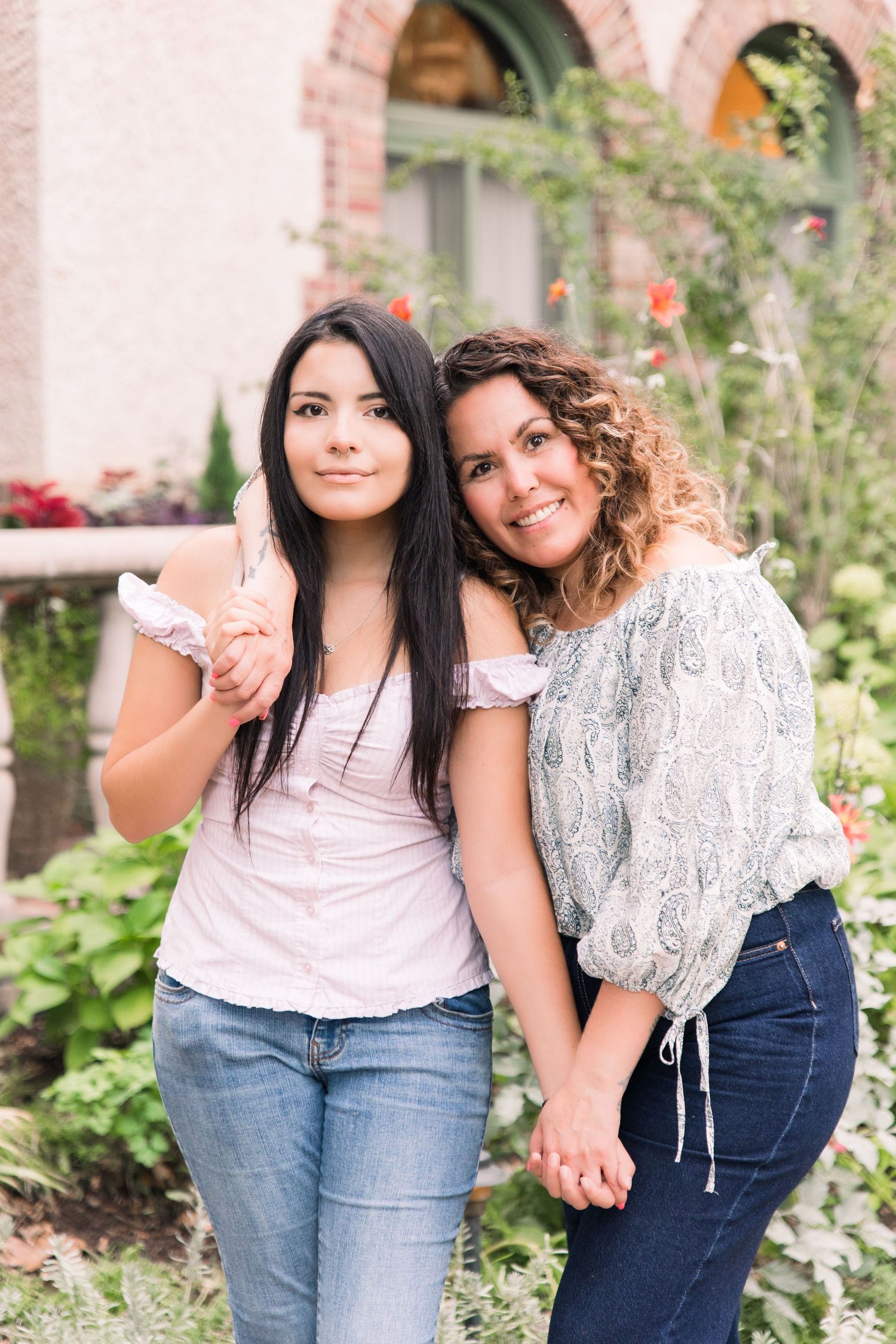 Two women smiling and posing together outdoors in a garden with colorful flowers and brick building backdrop.