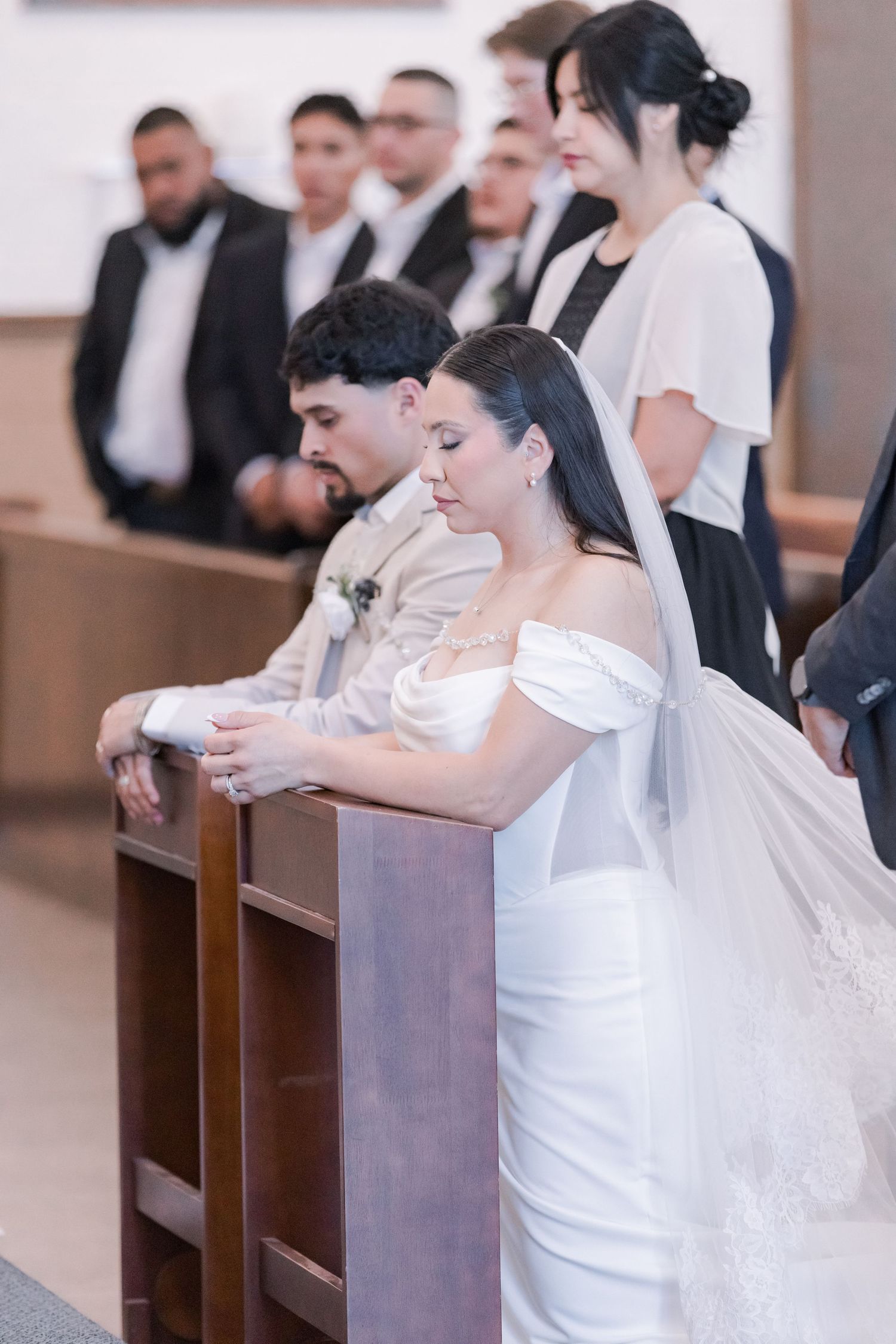 Bride in white veil and groom pray together at church pew during wedding ceremony.