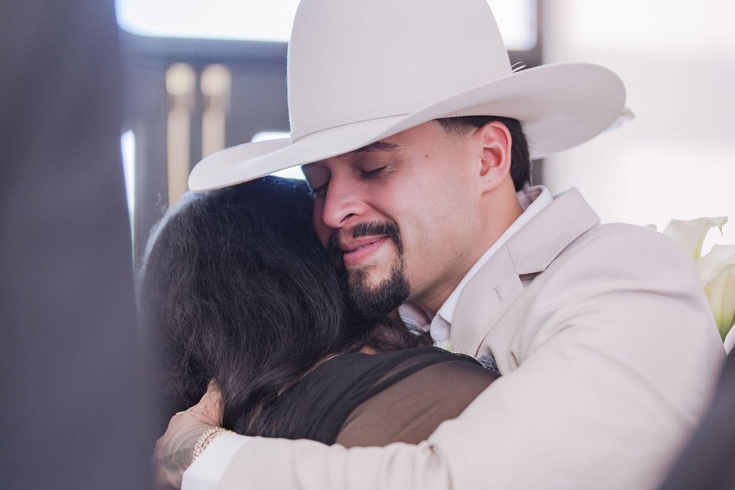 Joyful groom in white cowboy hat embraces a guest warmly during wedding reception celebration.