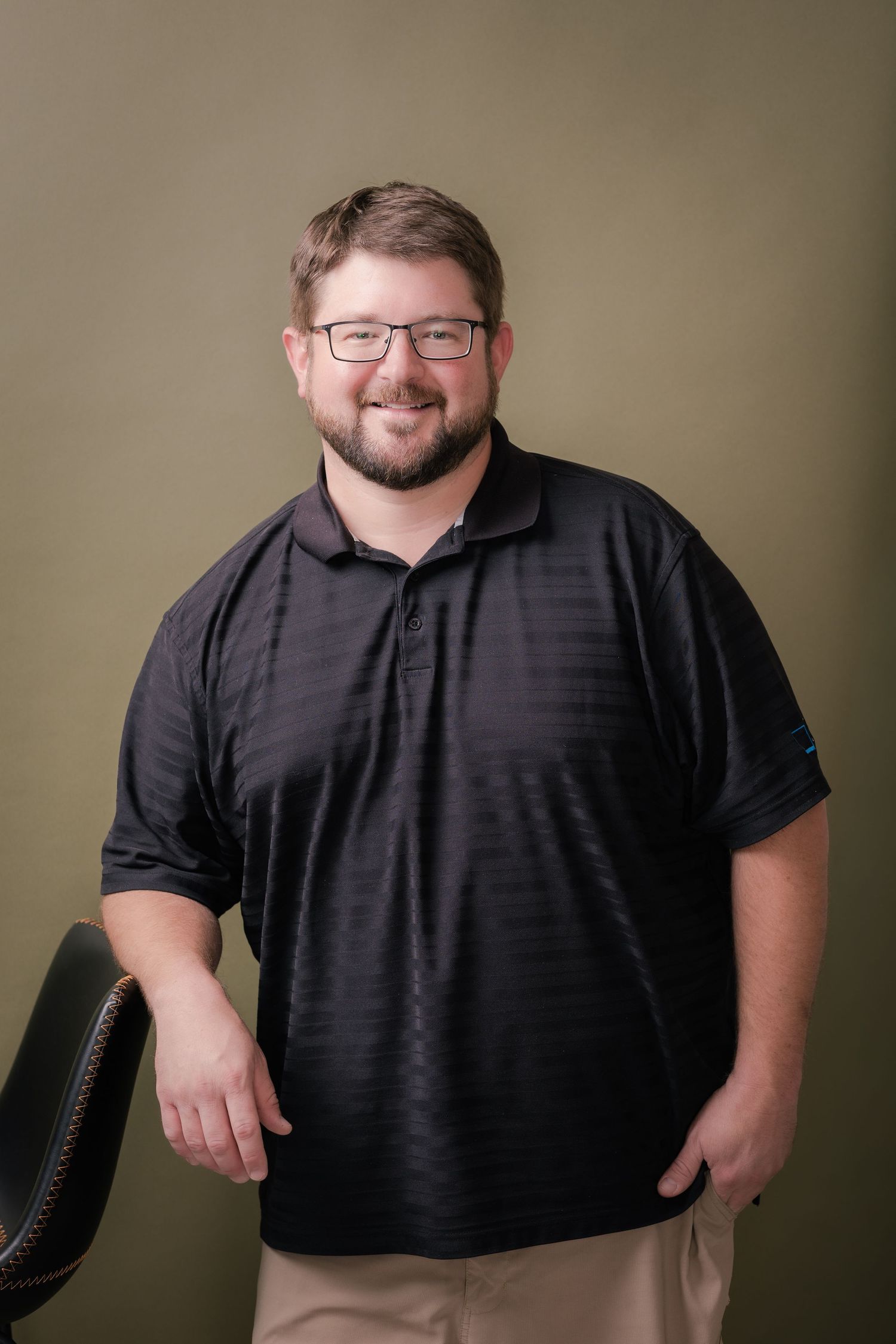 Smiling man with glasses and a beard wearing a black polo shirt and khaki pants, leaning on a chair against an olive green backdrop.