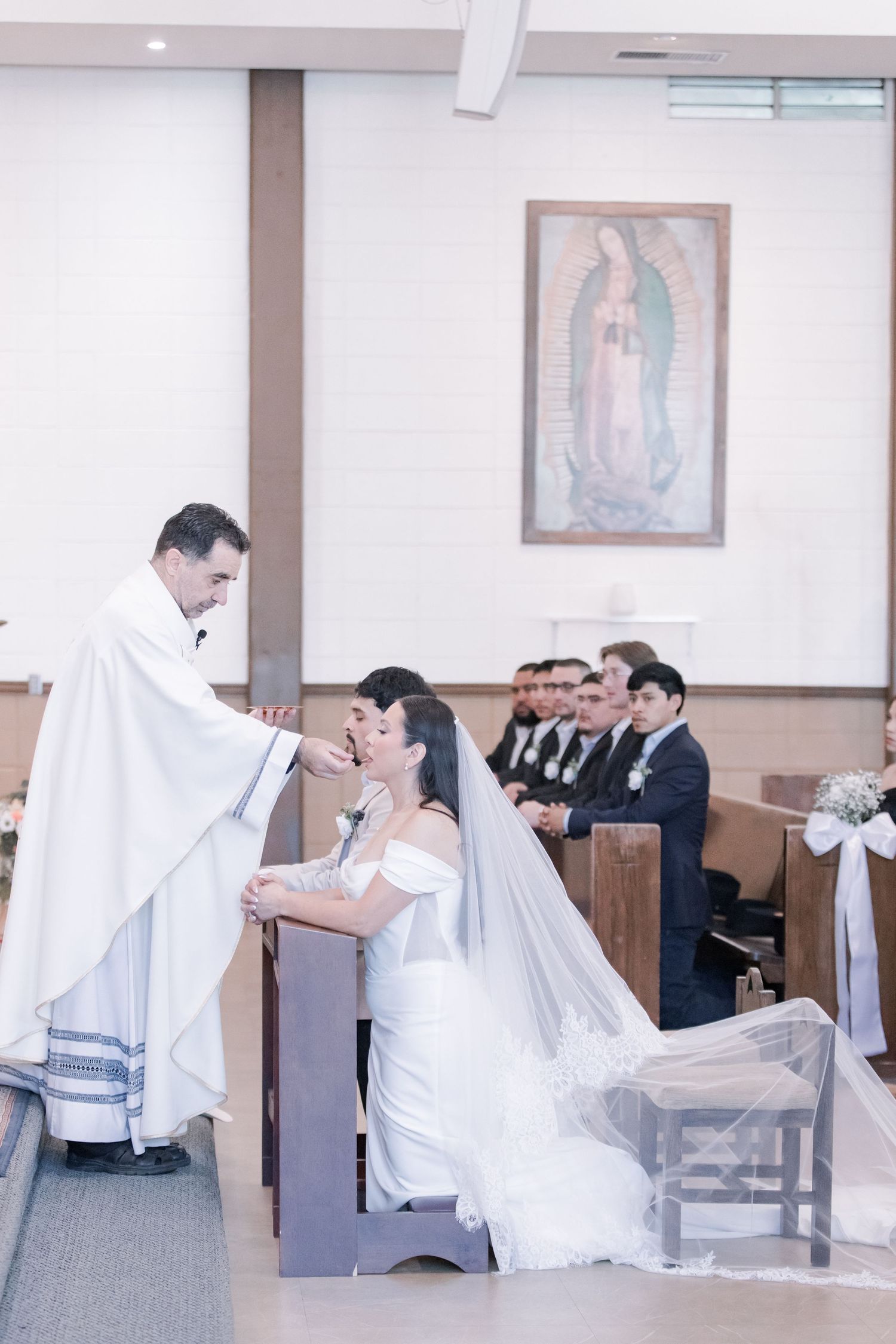 Priest places wedding crown on bride during Catholic ceremony with Our Lady of Guadalupe image behind.