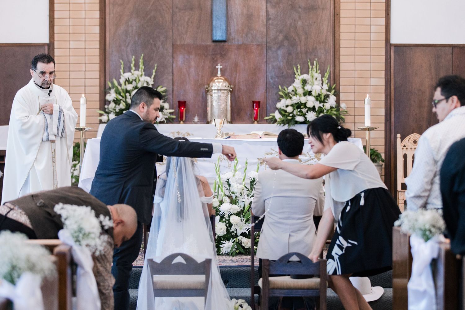 Wedding party assists bride and groom at church altar during Catholic wedding ceremony ritual.