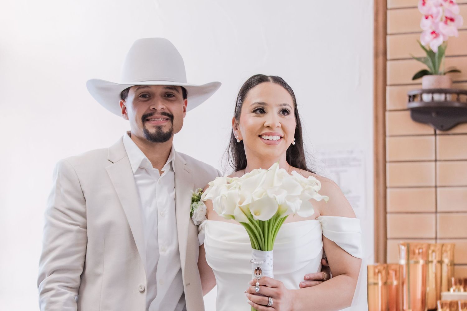 Smiling newlyweds pose together, groom wearing white cowboy hat, bride holding white calla lily bouquet.