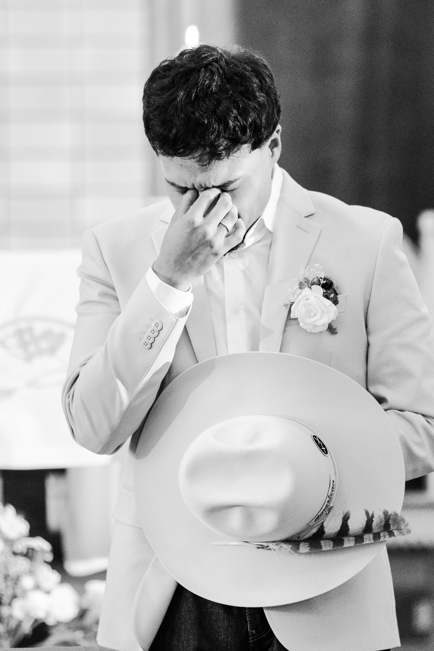 Emotional groom in beige suit wipes tears holding cowboy hat at wedding ceremony, black and white photo.