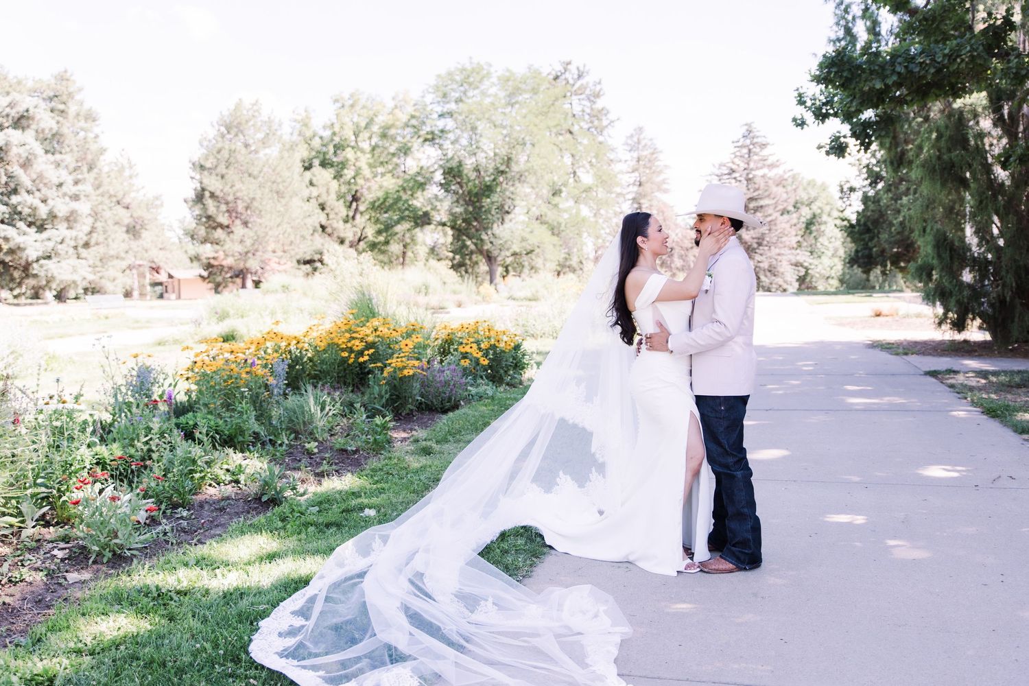 Bride in flowing white gown and groom in pink suit share romantic moment on garden path with colorful flowers.