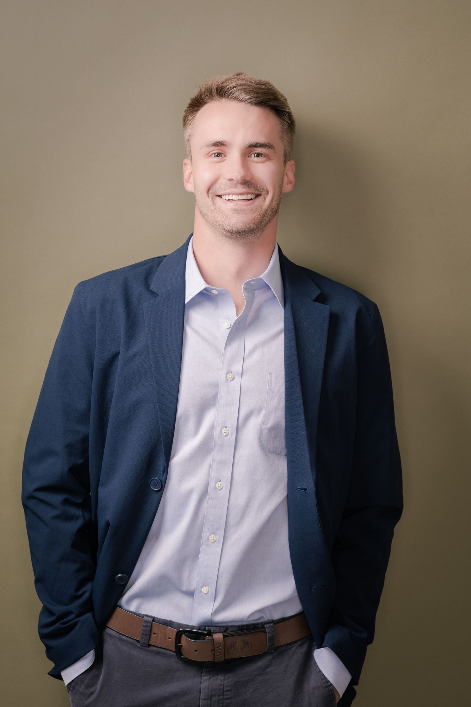Smiling man in a navy blazer and light blue button-up shirt stands with hands in pockets against an olive green wall.