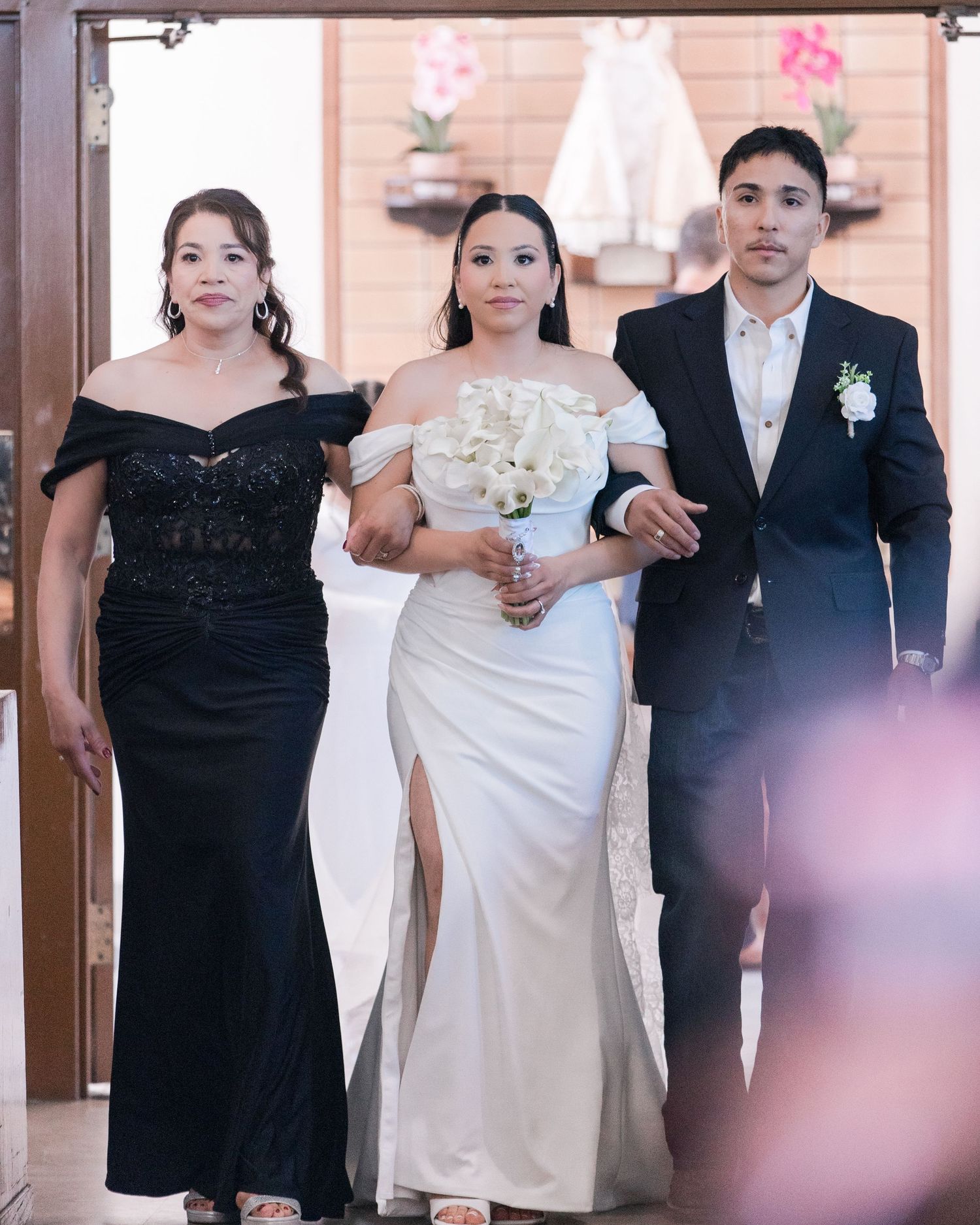 Bride in white off-shoulder wedding dress walks down aisle escorted by both parents.
