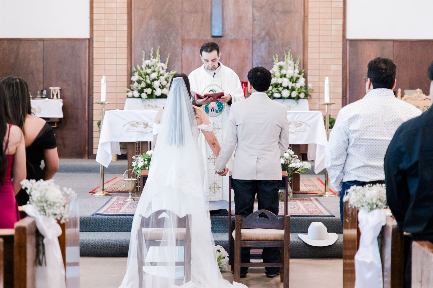 Bride and groom hold hands before priest at decorated church altar during wedding ceremony.