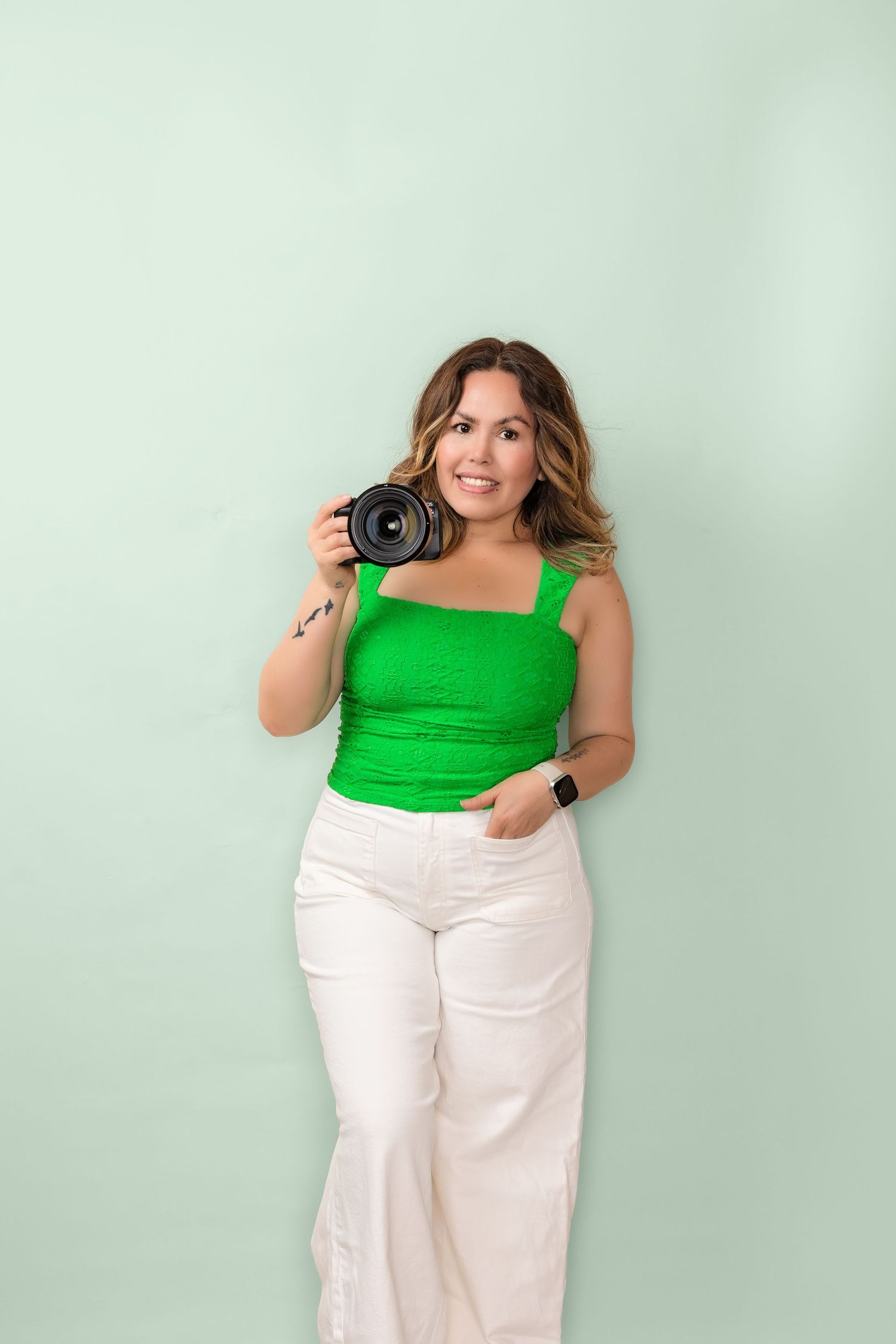 Woman in green top and white jeans holding a camera, smiling against a mint green background.
