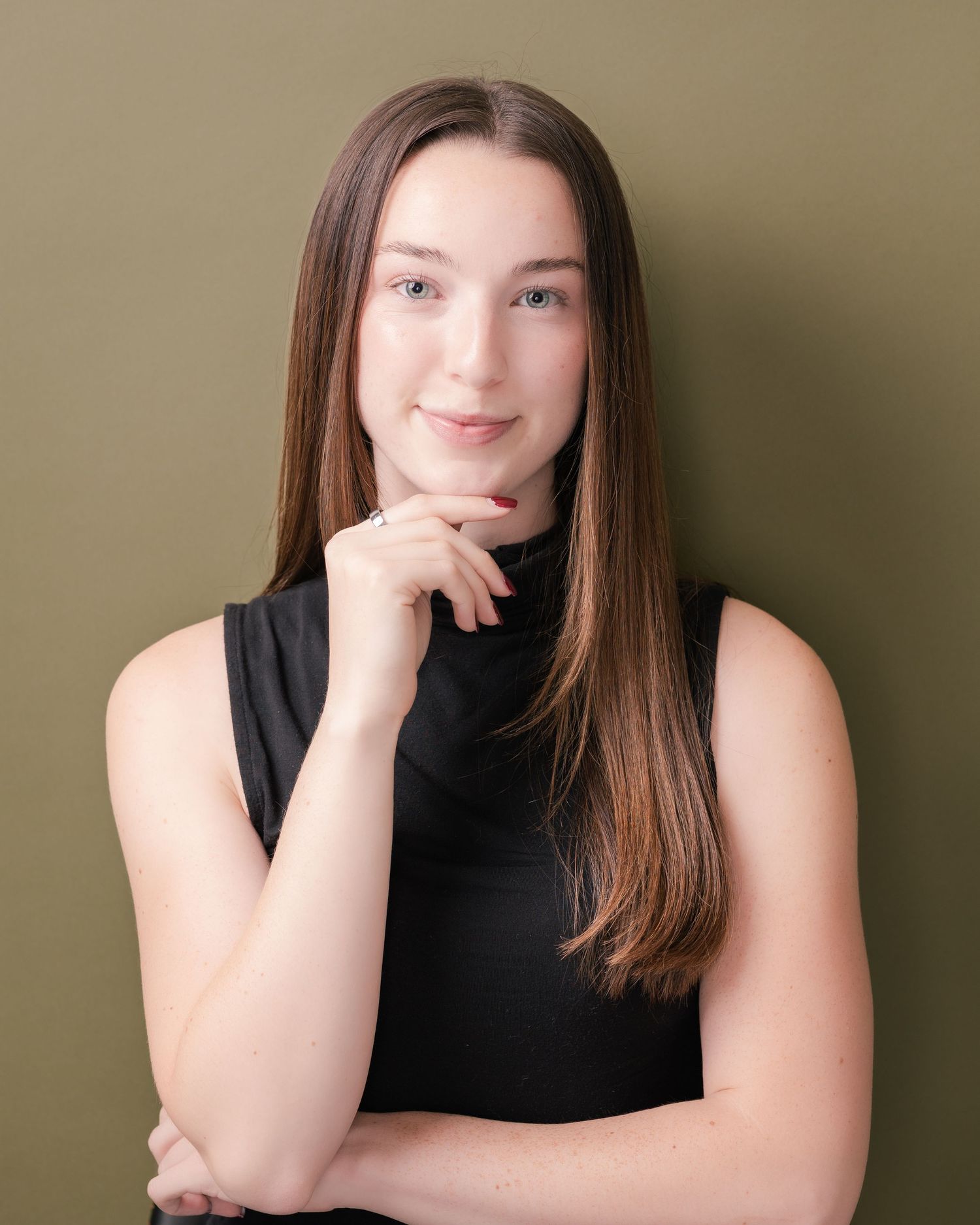 Young woman with long straight brown hair in a black sleeveless turtleneck poses with her hand on her chin against an olive green backdrop.