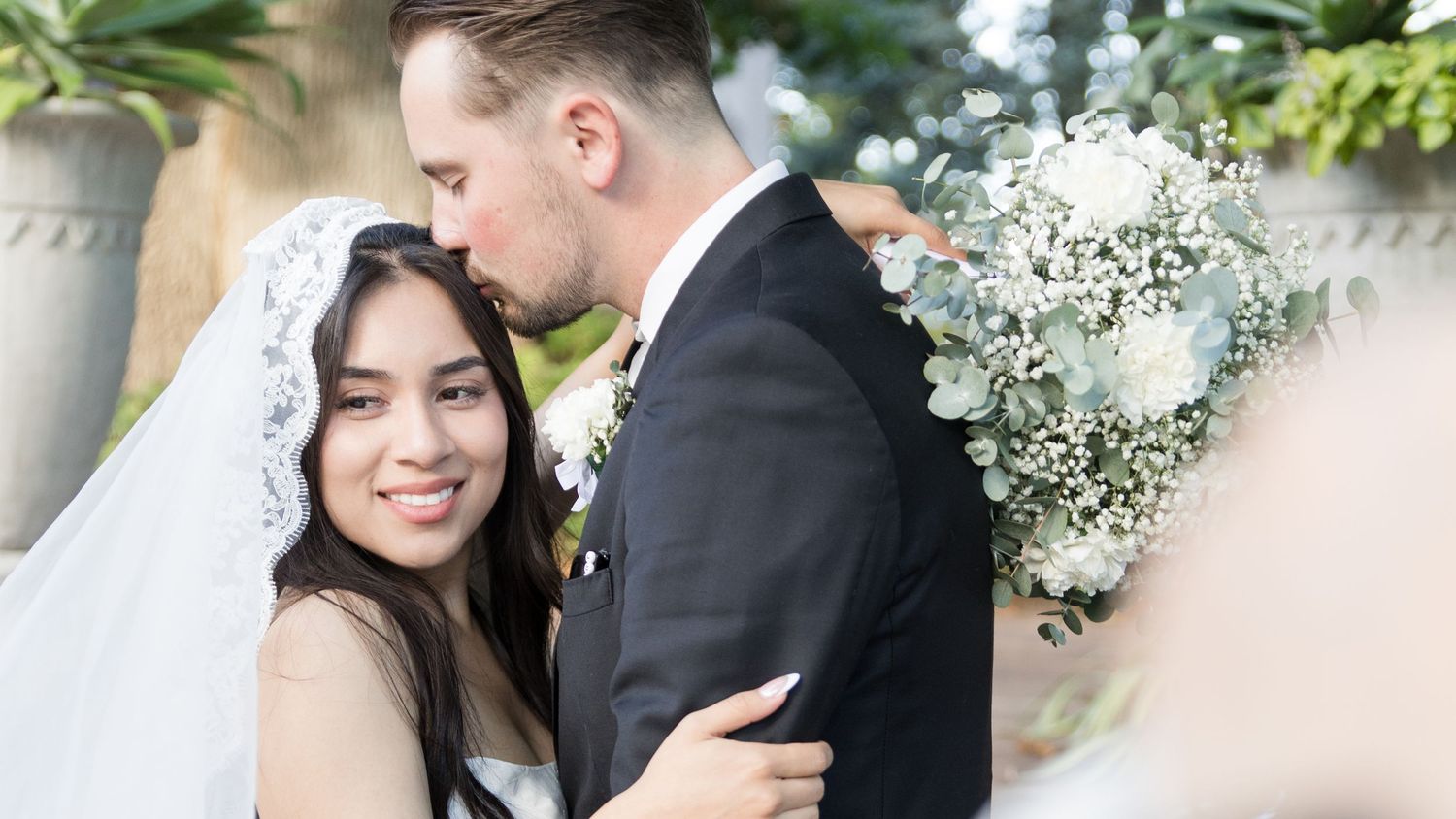 Groom in a dark suit kisses the forehead of a smiling bride in a lace veil as she holds a white floral bouquet at an outdoor wedding.