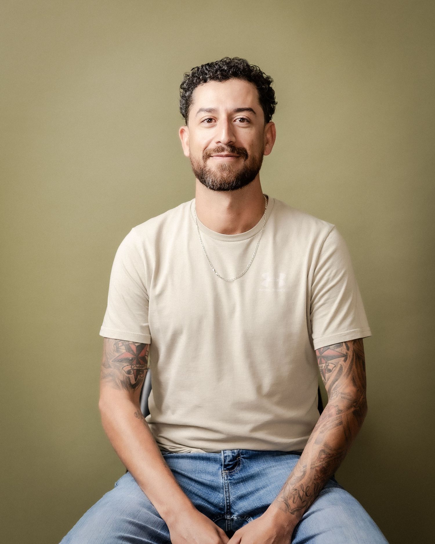 Smiling man with curly dark hair, beard and tattooed forearms sits in a cream t-shirt and jeans against an olive green backdrop.