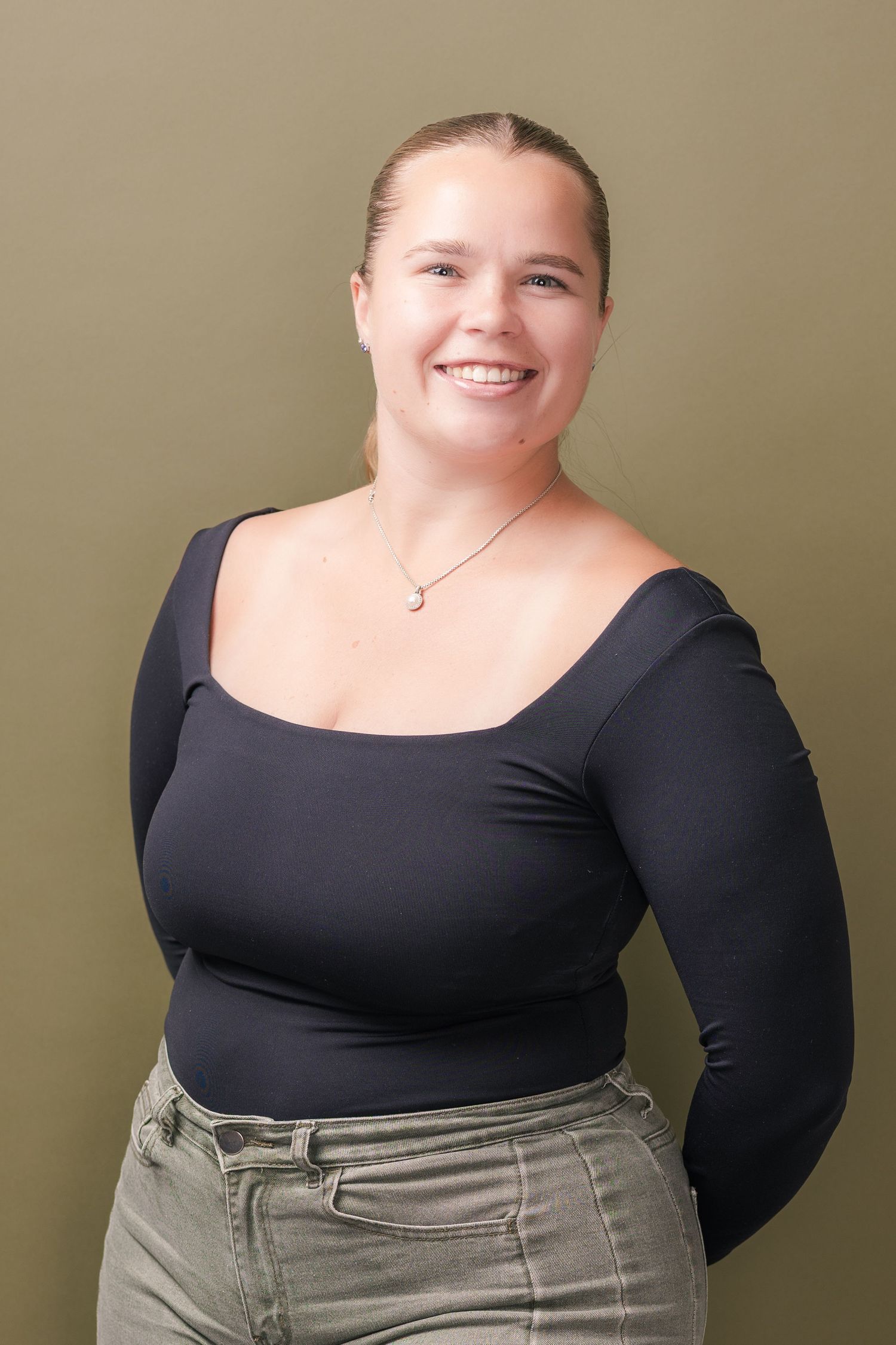 Smiling woman with hair pulled back wears a black square-neck long-sleeve top, pendant necklace and olive pants against an olive backdrop.