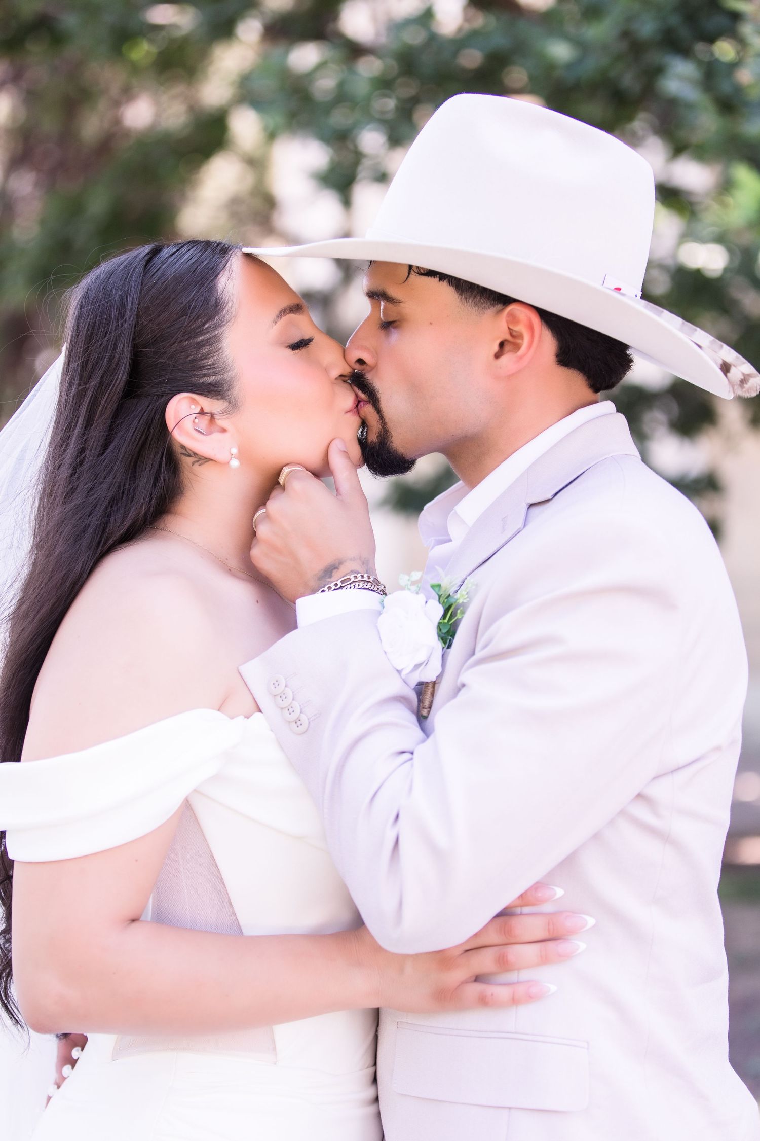 Bride and groom share a romantic kiss outdoors, groom in tan suit and white cowboy hat.