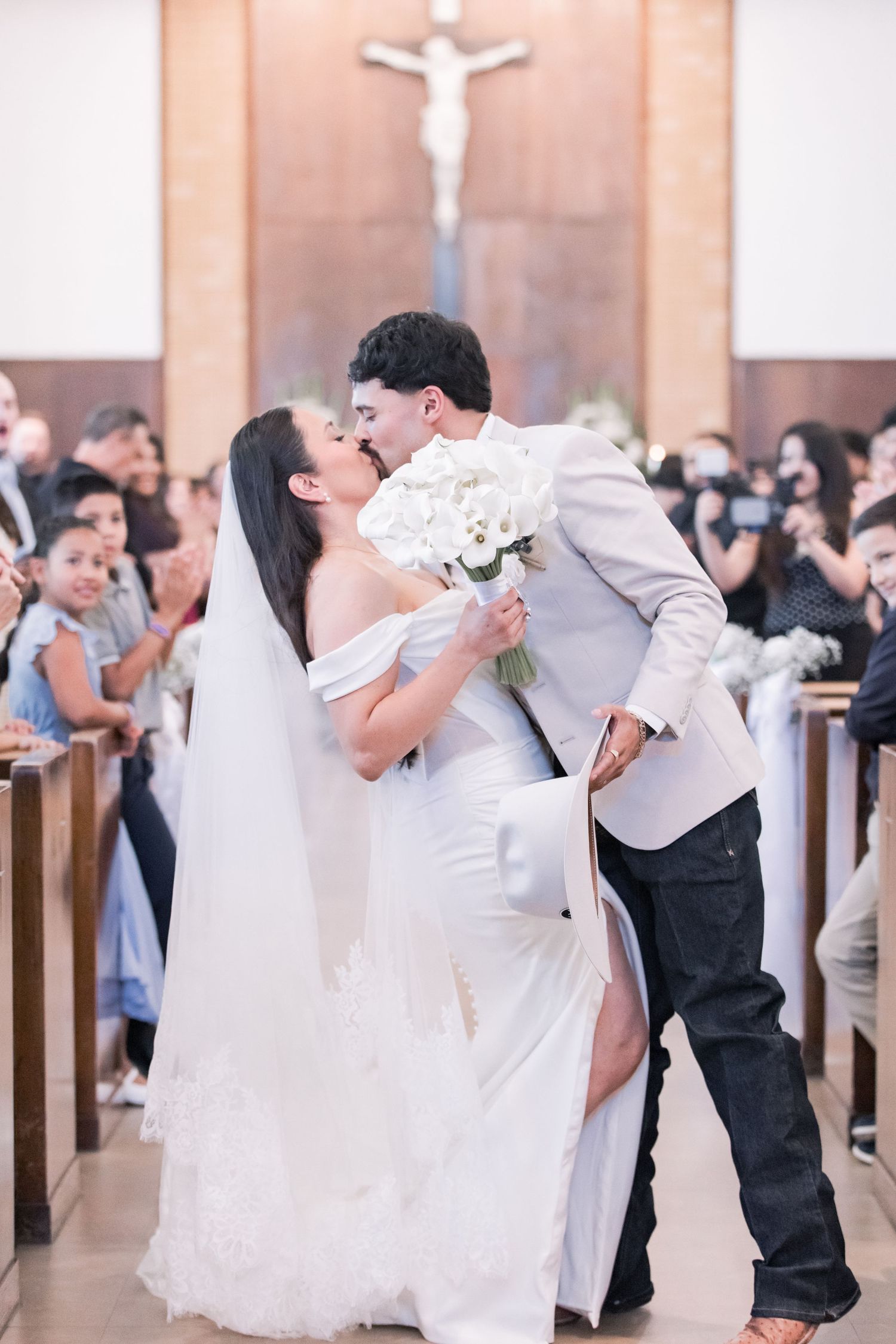Newlyweds share first kiss at church altar as guests celebrate, bride holding white bouquet.