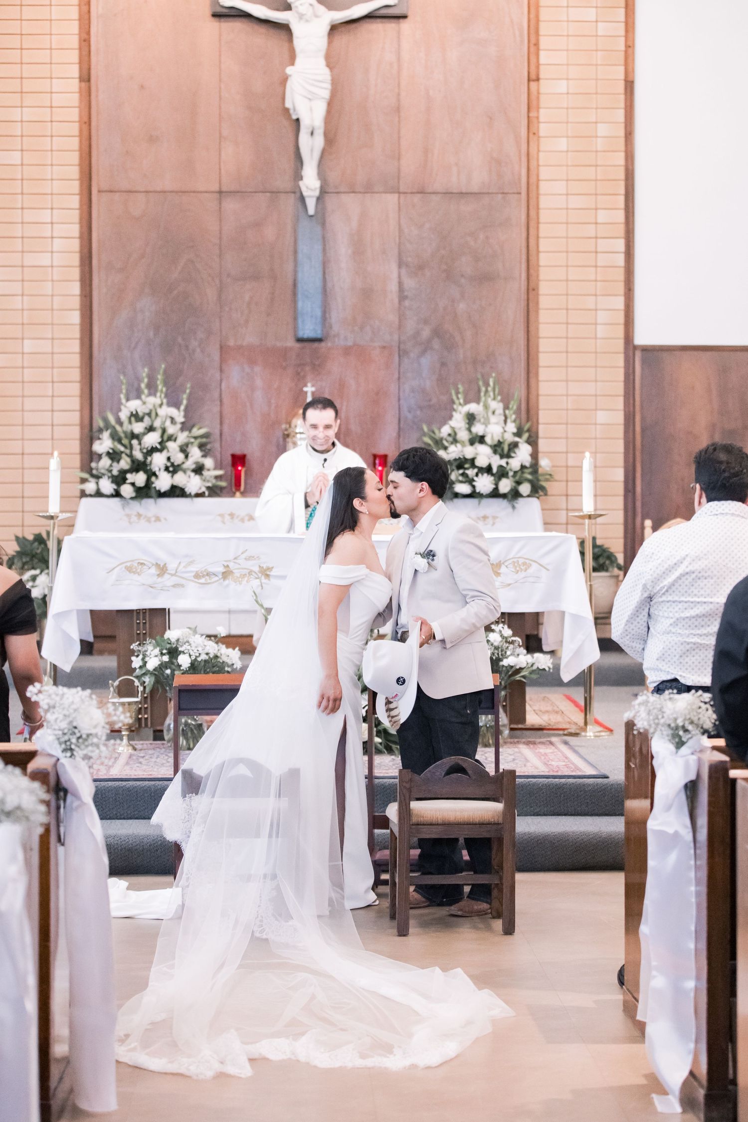 Bride and groom share first kiss at Catholic church altar during wedding ceremony with priest officiating.