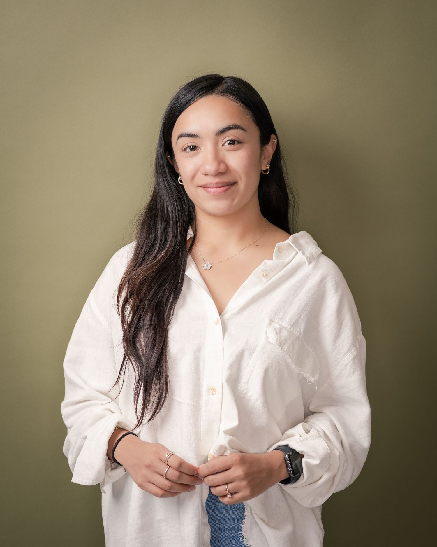 Smiling young woman with long dark hair in a white button-up shirt and jeans poses against an olive green background.