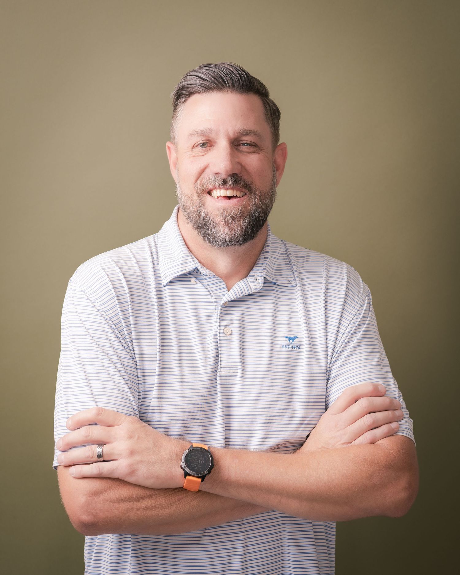 Smiling bearded man with arms crossed wearing a striped polo shirt and smartwatch against an olive green studio background.