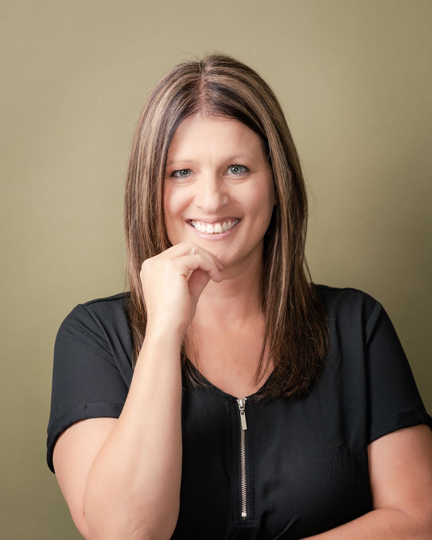 Smiling woman with long brown highlighted hair and a black zip-front top rests her chin on her hand against an olive green background.