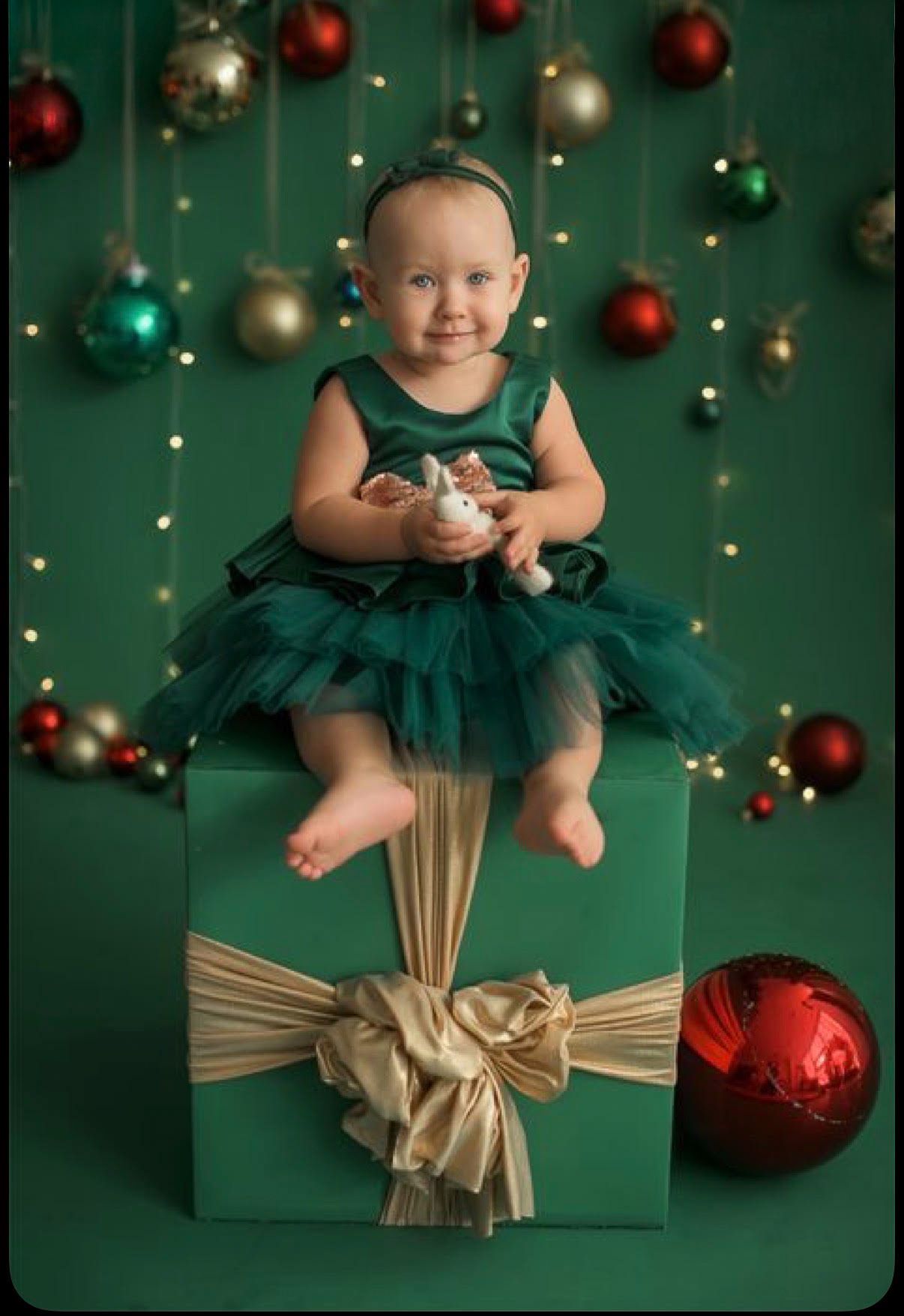 Baby girl in a green tulle dress sits on a green gift box with gold ribbon, surrounded by hanging Christmas ornaments and twinkling lights.