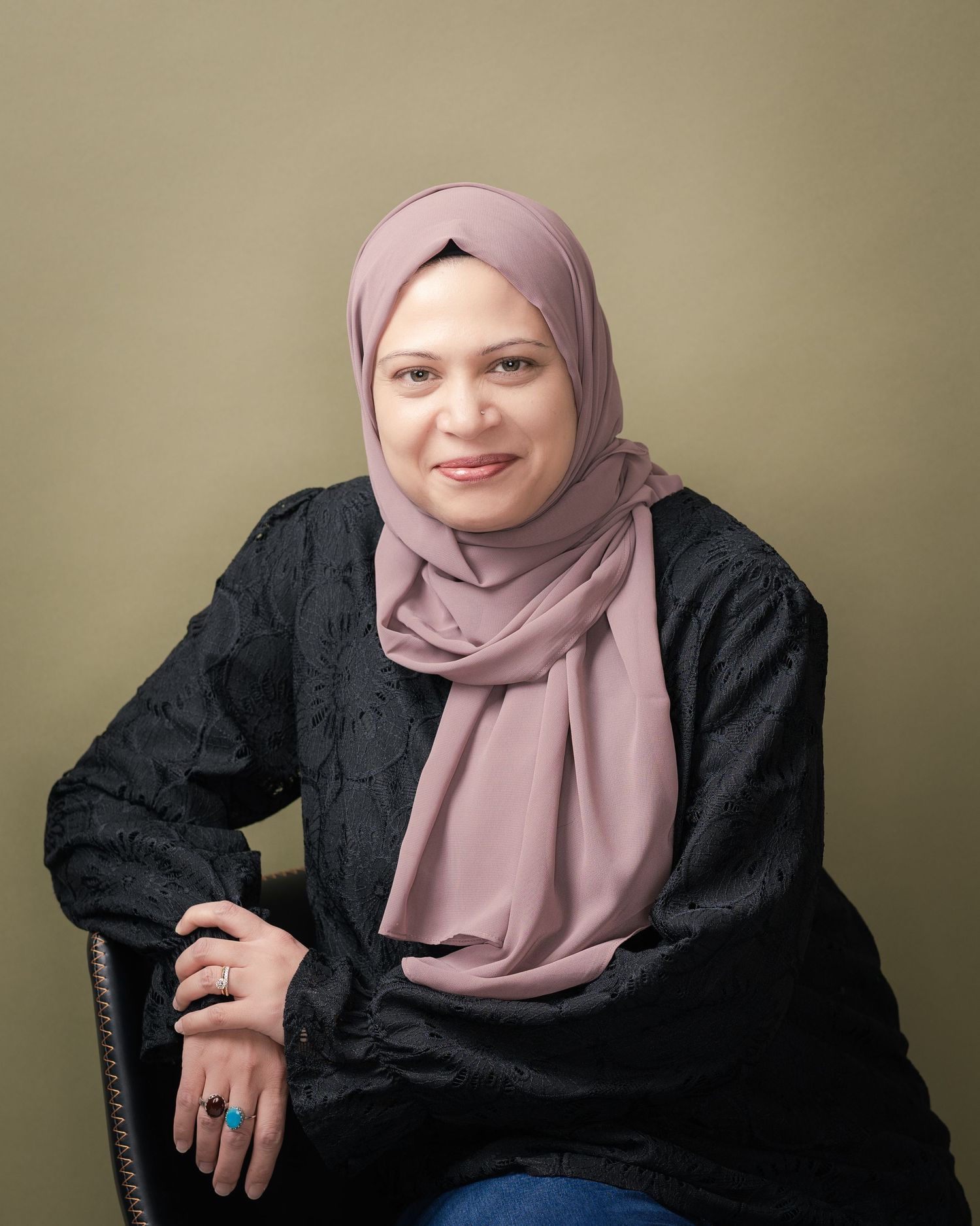 Smiling woman in a mauve hijab and black embroidered top sits in a chair against an olive green backdrop, wearing colorful rings.