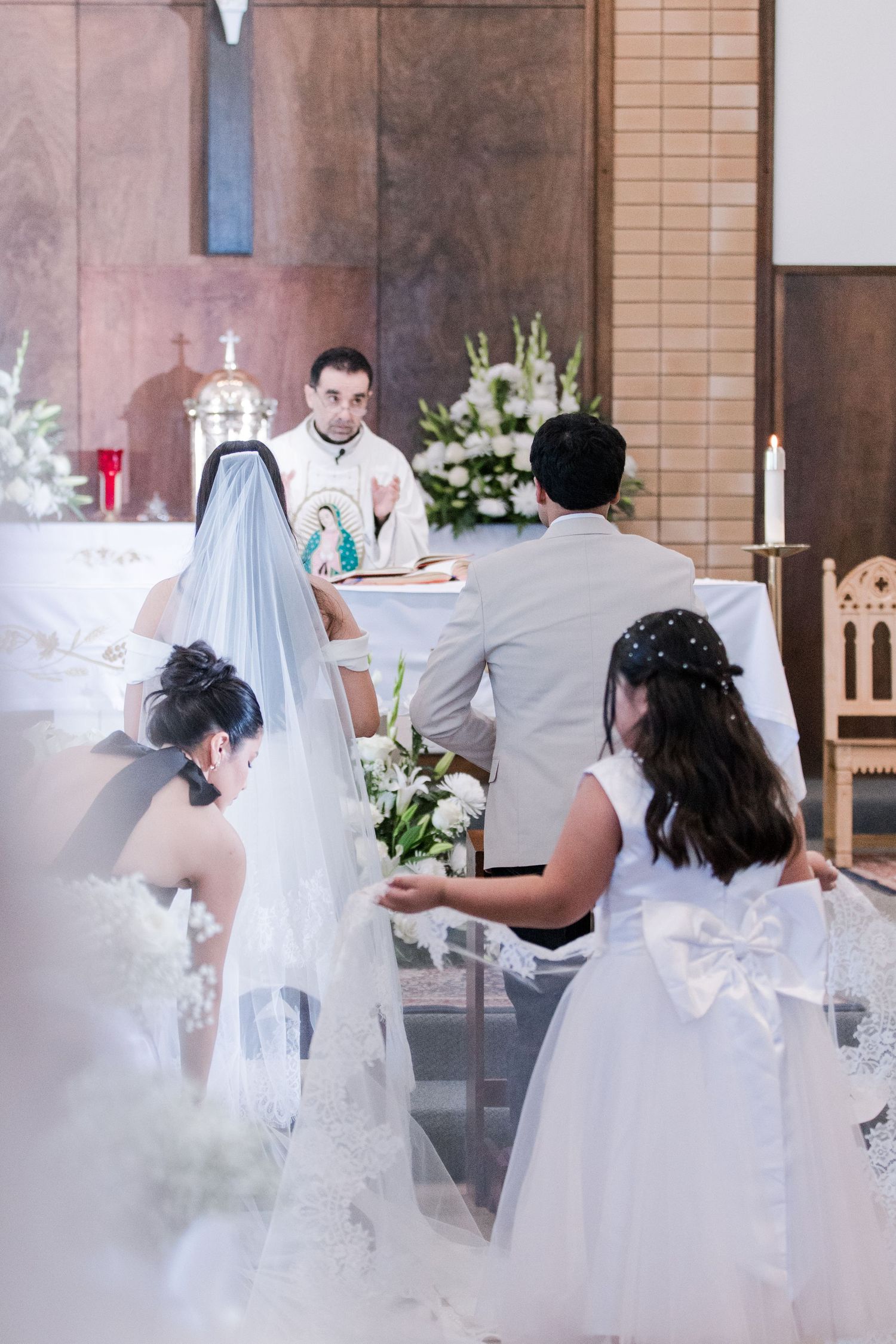 Priest reads scripture as bride and groom kneel at altar during Catholic wedding ceremony.