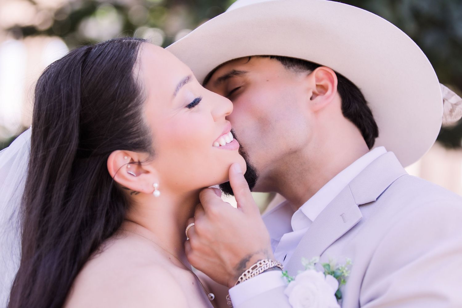 Close-up of groom in cowboy hat kissing smiling bride on cheek at outdoor wedding.