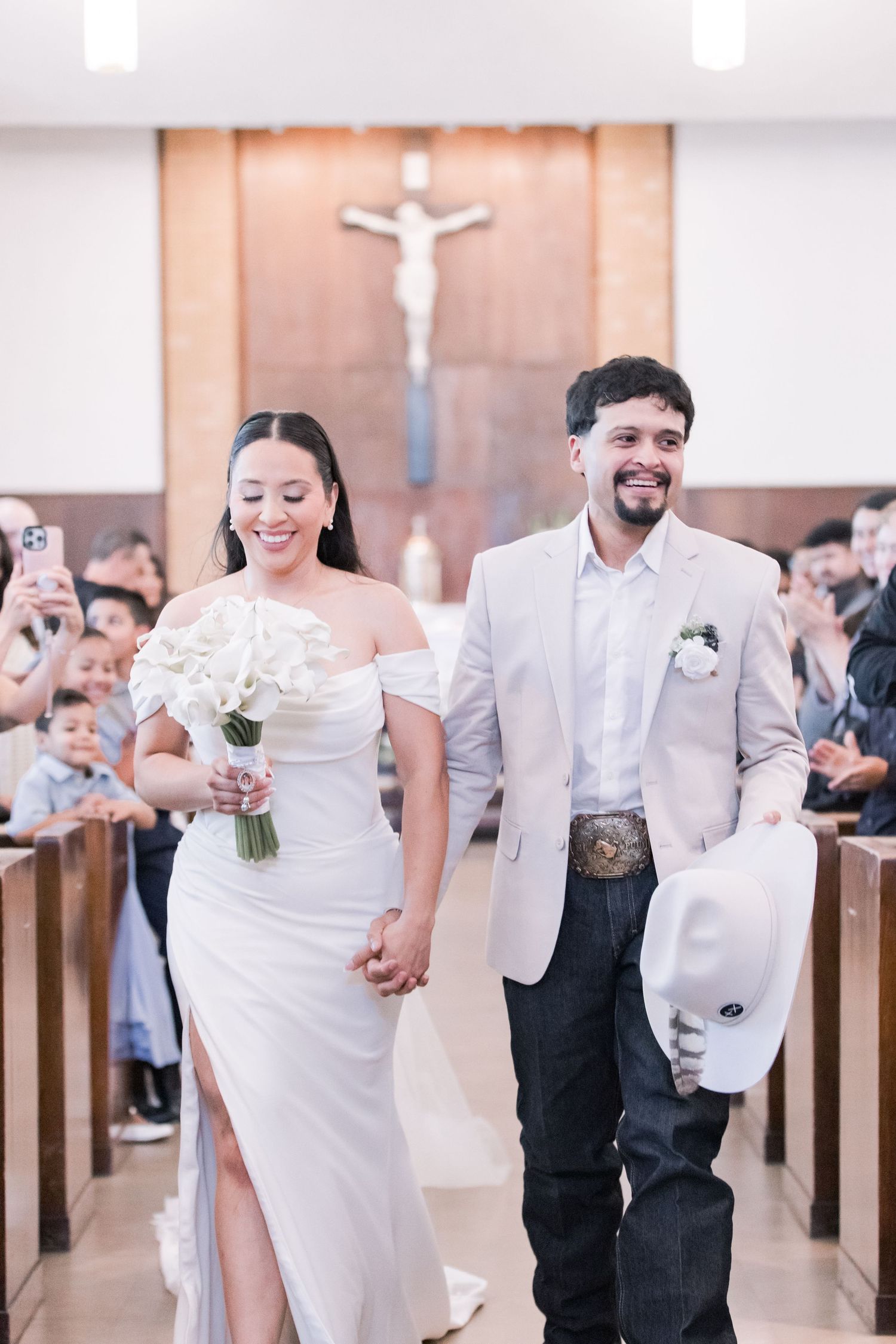 Happy newlyweds walk down church aisle smiling, bride holding white bouquet, groom carrying cowboy hat.