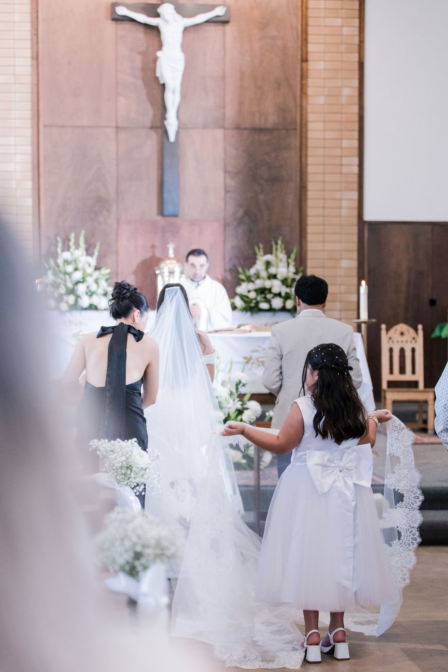 Bride at Catholic church altar with flower girl holding veil train during wedding ceremony.