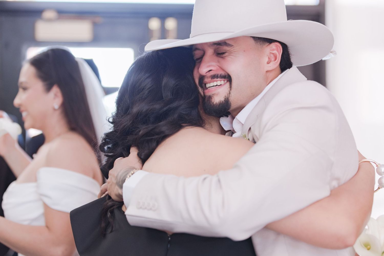 Beaming groom in white cowboy hat hugs a wedding guest while bride stands nearby in background.