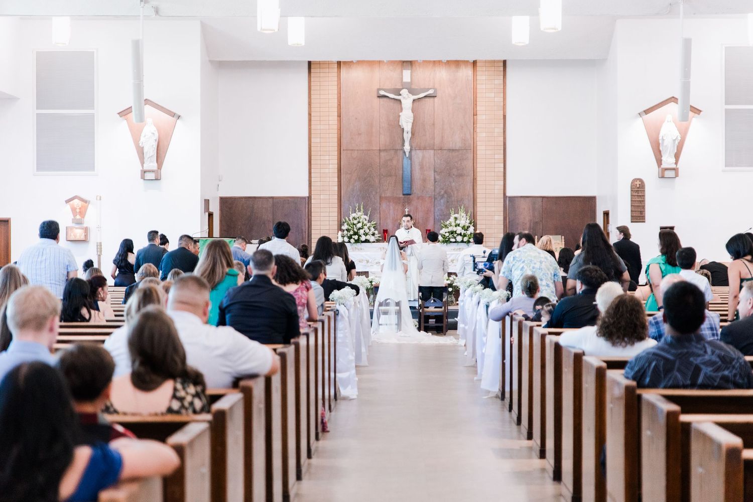 Wide view of Catholic church filled with wedding guests during ceremony with couple at altar.