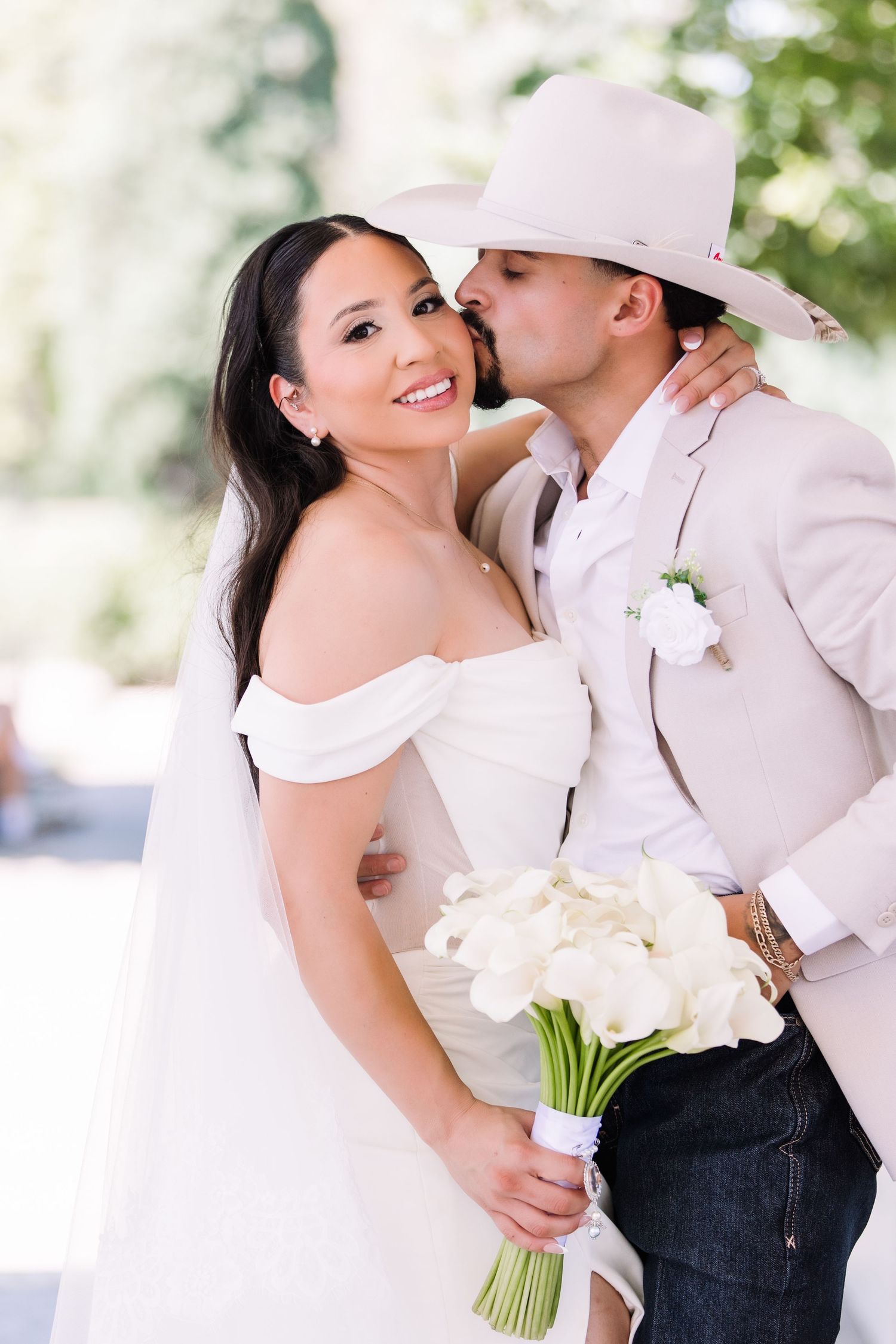 Bride holding white calla lily bouquet smiles as groom in tan cowboy hat and suit kisses her cheek.
