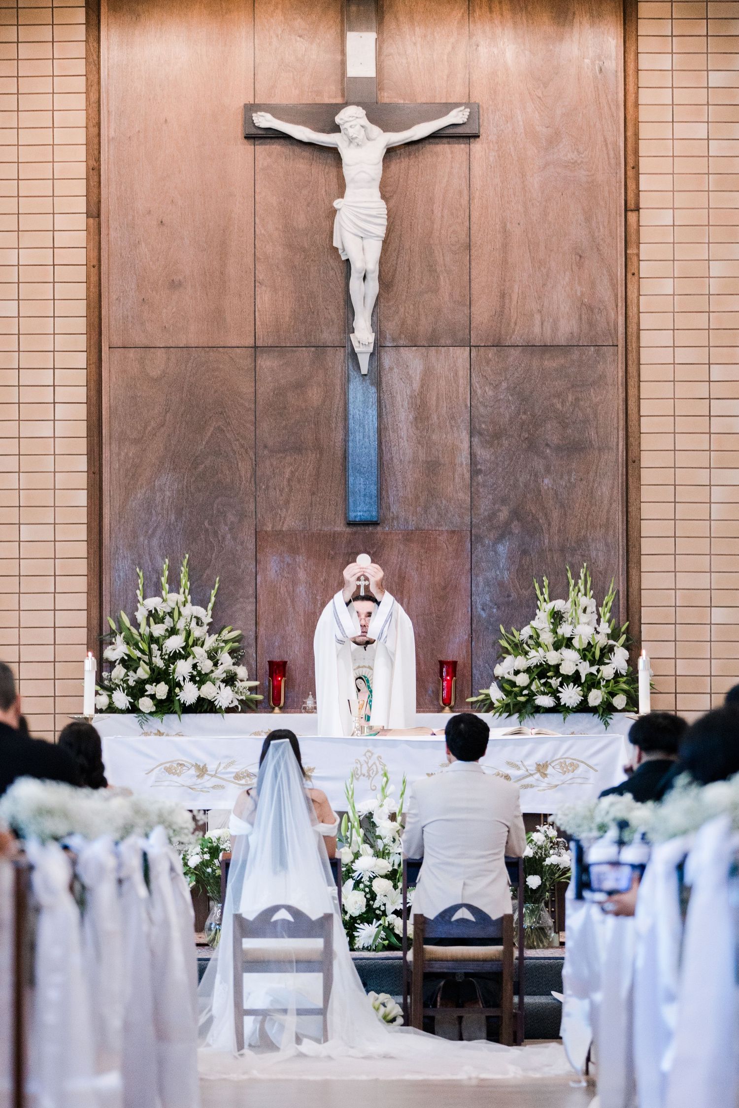 Bride and groom kneel before priest at decorated altar with crucifix during Catholic wedding Mass.