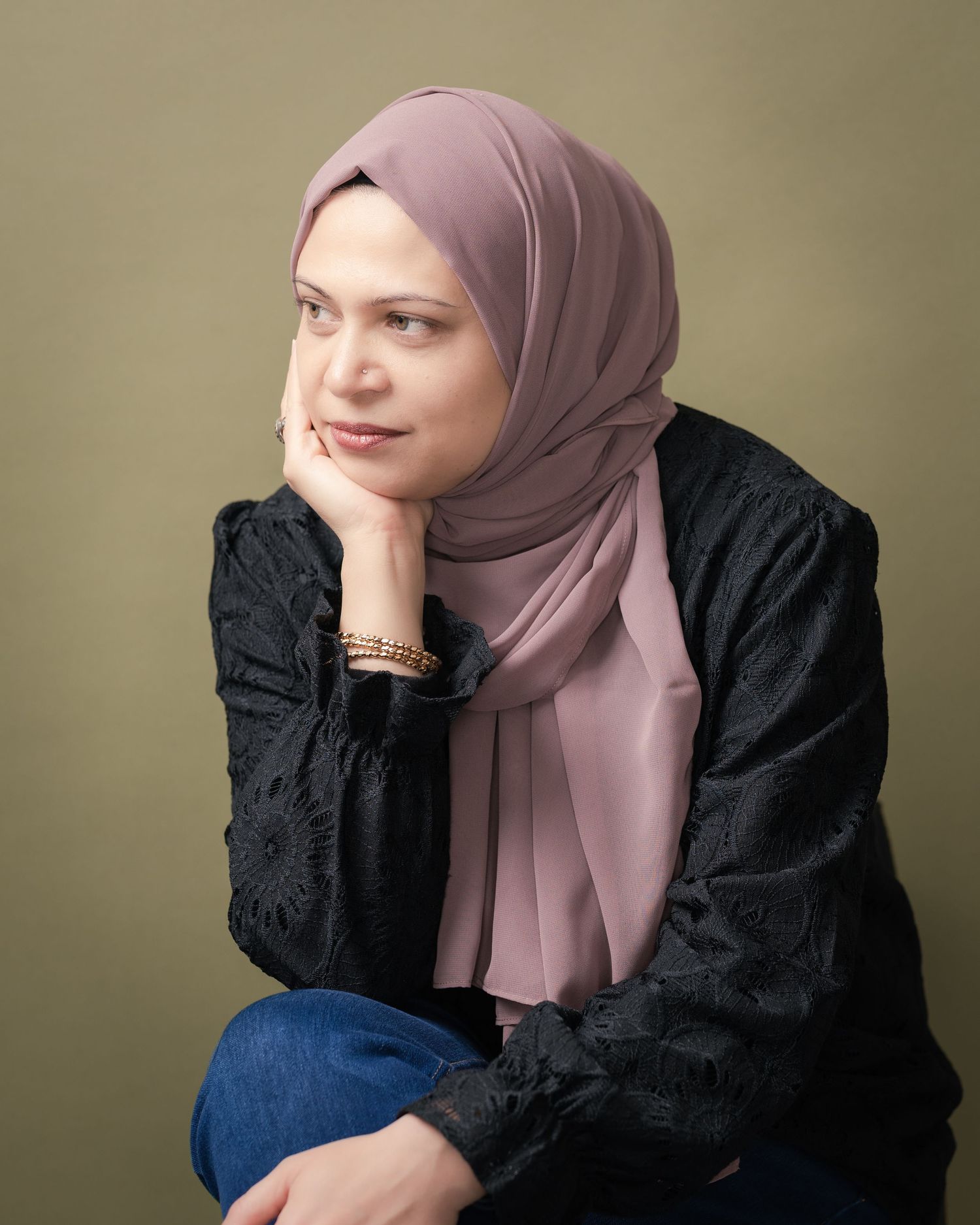 Woman in a dusty rose hijab and black embroidered blouse rests her chin on her hand, gazing thoughtfully against an olive green backdrop.
