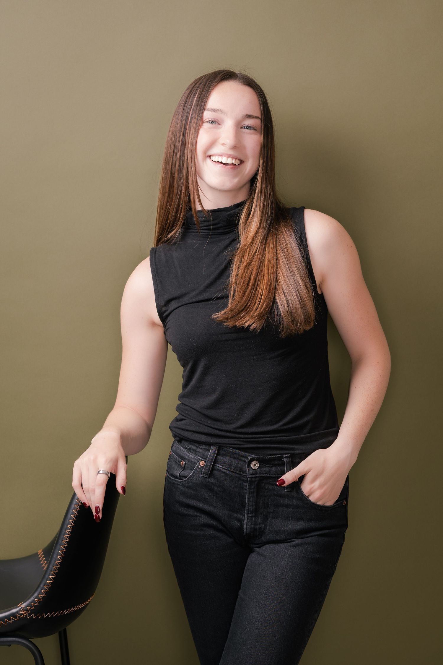 Smiling woman with long brown hair in a black sleeveless mock-neck top and black jeans leans on a black chair against an olive green wall.